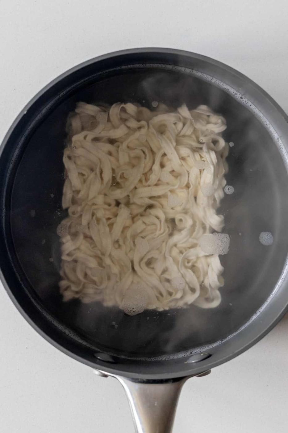 A black pot of boiling water with uncooked, flat, white noodles being cooked, perfect as a base for a flavorful Birria Ramen.