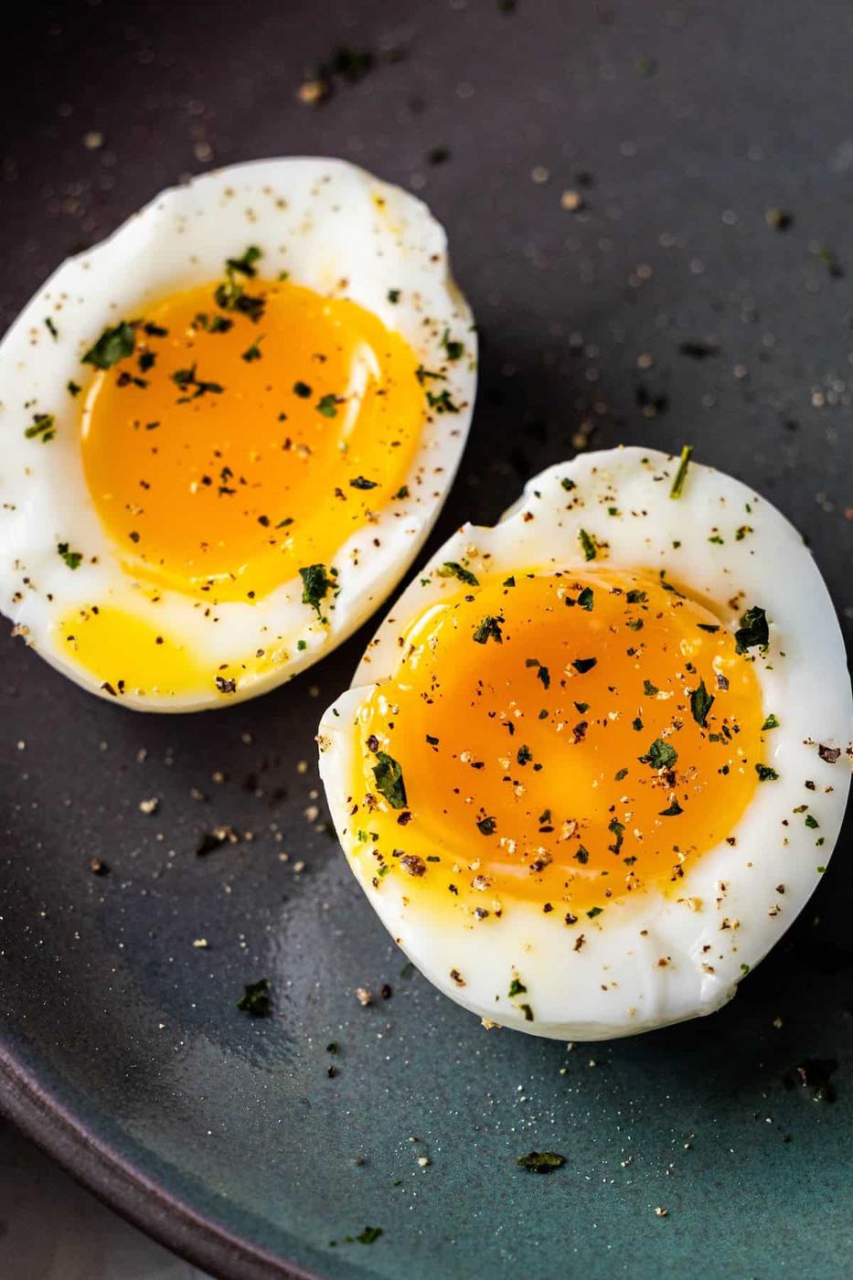 Two soft-boiled egg halves with runny yolks, sprinkled with black pepper and herbs, on a dark plate—perfect as a savory topping for your favorite bowl of ramen.