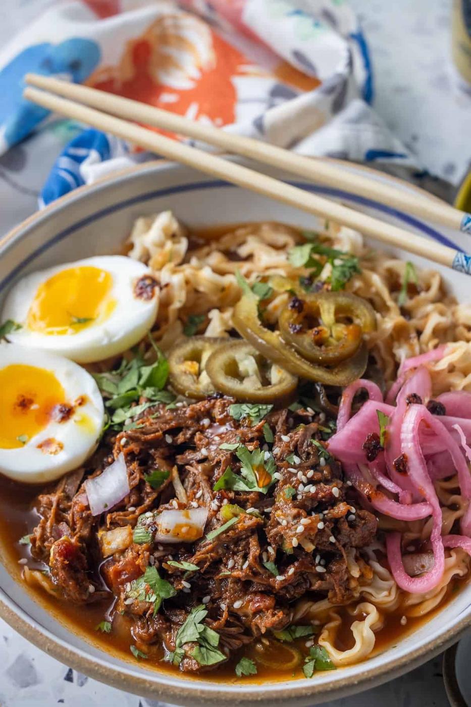 Bowl of Birria Ramen noodles topped with shredded meat, soft-boiled eggs, pickled onions, jalapeños, chopped herbs, and sesame seeds, with chopsticks resting on the bowl’s rim.