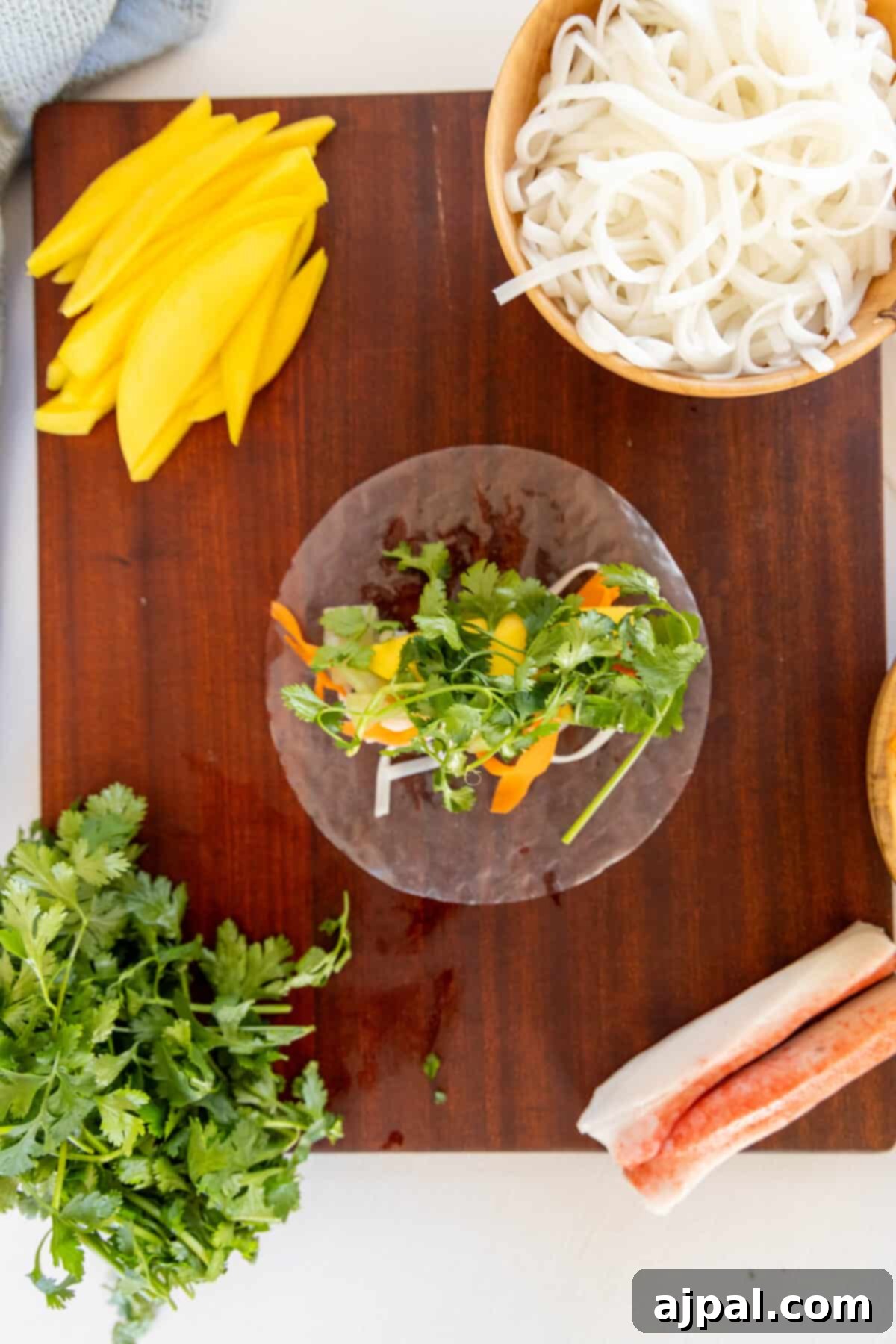 A wooden cutting board with ingredients for a Thai noodle dish and crab spring rolls.