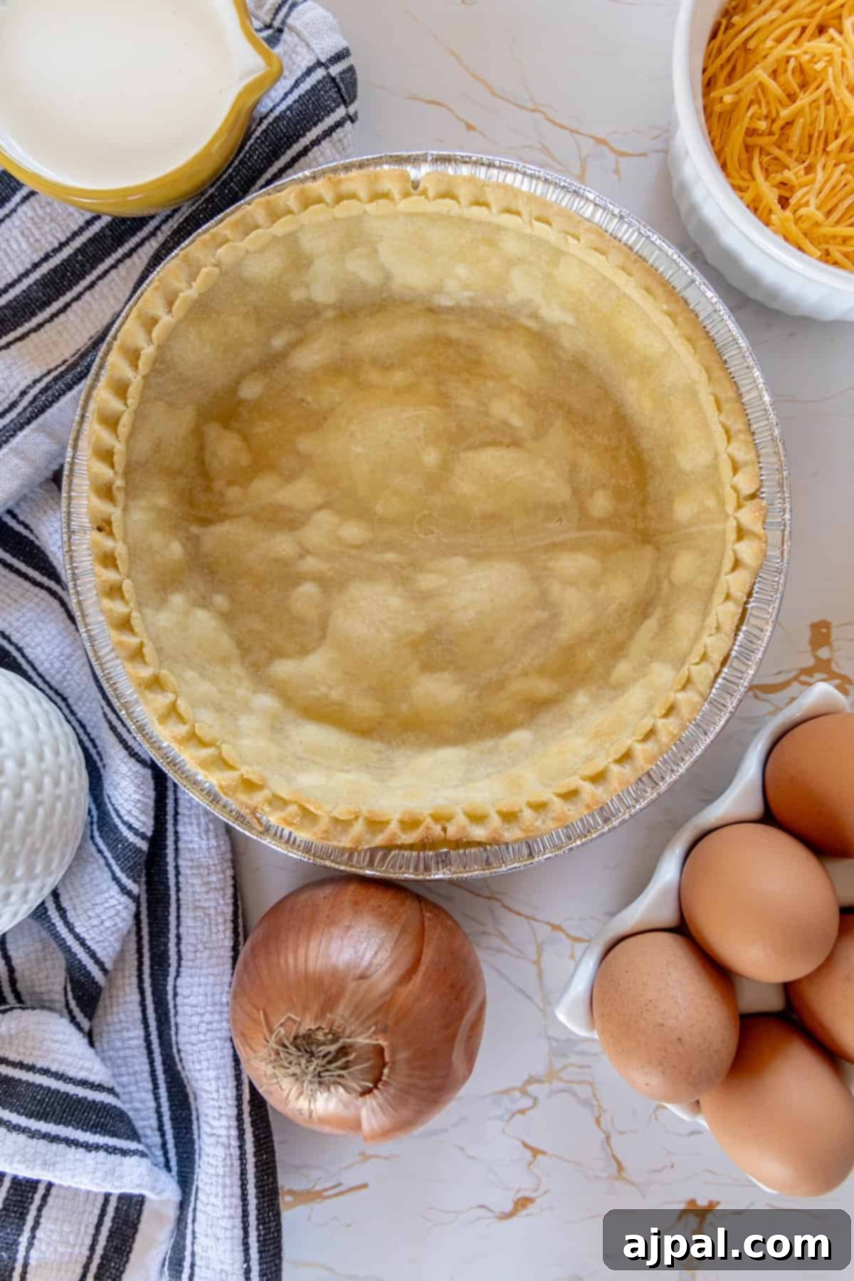 A pie pan filled with a quiche crust, surrounded by raw eggs and chopped onions, ingredients for the filling.