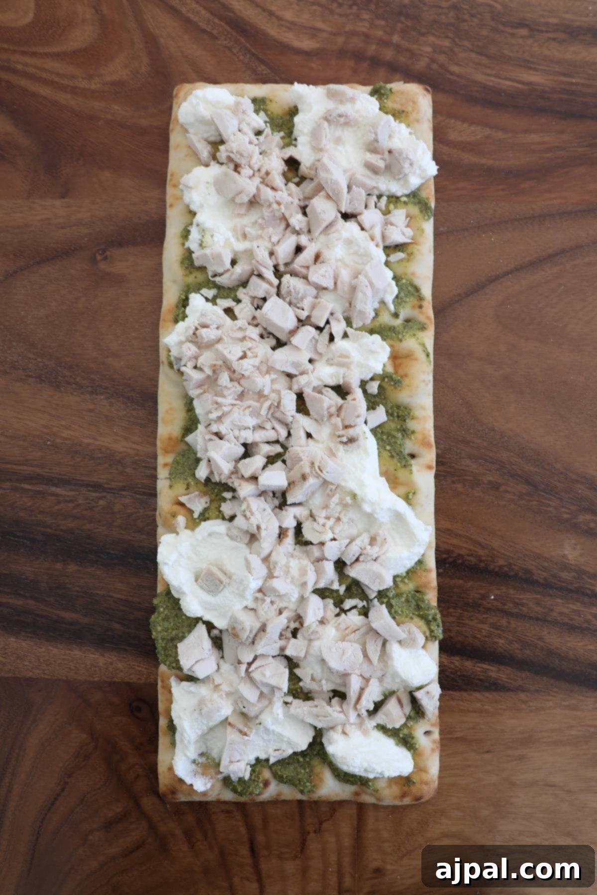 Artisan Pesto Chicken Flatbread 7 A partially assembled flatbread pizza showcasing cooked chicken pieces and dollops of creamy white cheese on a pesto base.