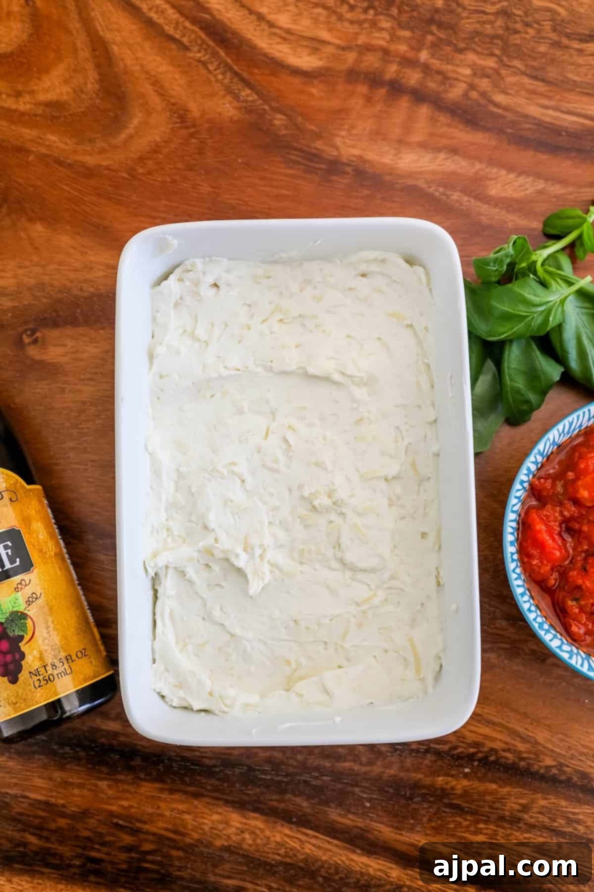 A pristine white dish holds a medley of fresh tomatoes, basil, and seasonings alongside a bowl of creamy ricotta cheese, poised for baking a savory bruschetta creation.