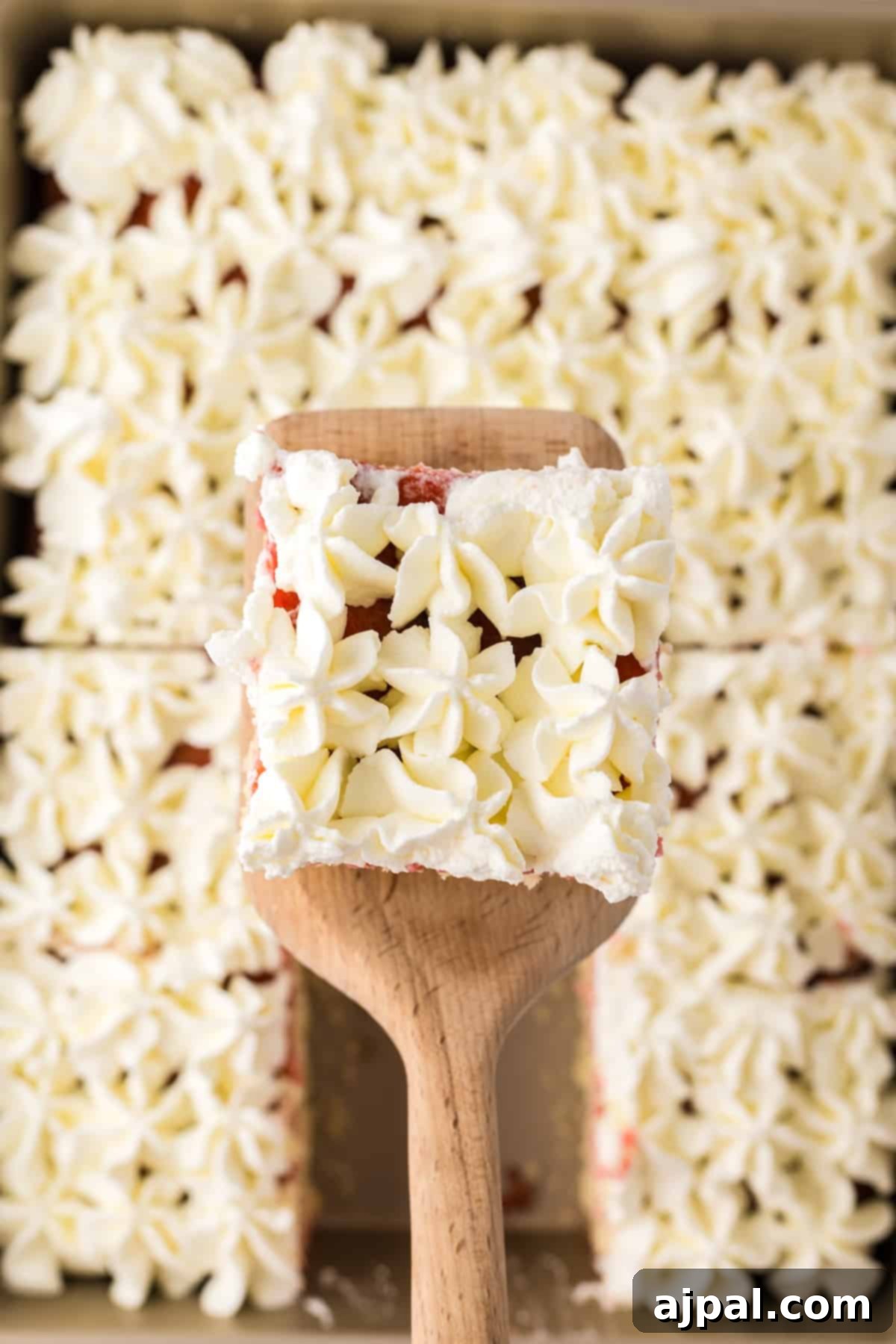 A square slice of raspberry sheet cake, showcasing its moist interior and generous frosting, sits in a baking pan beside a wooden spatula, ready to be served.