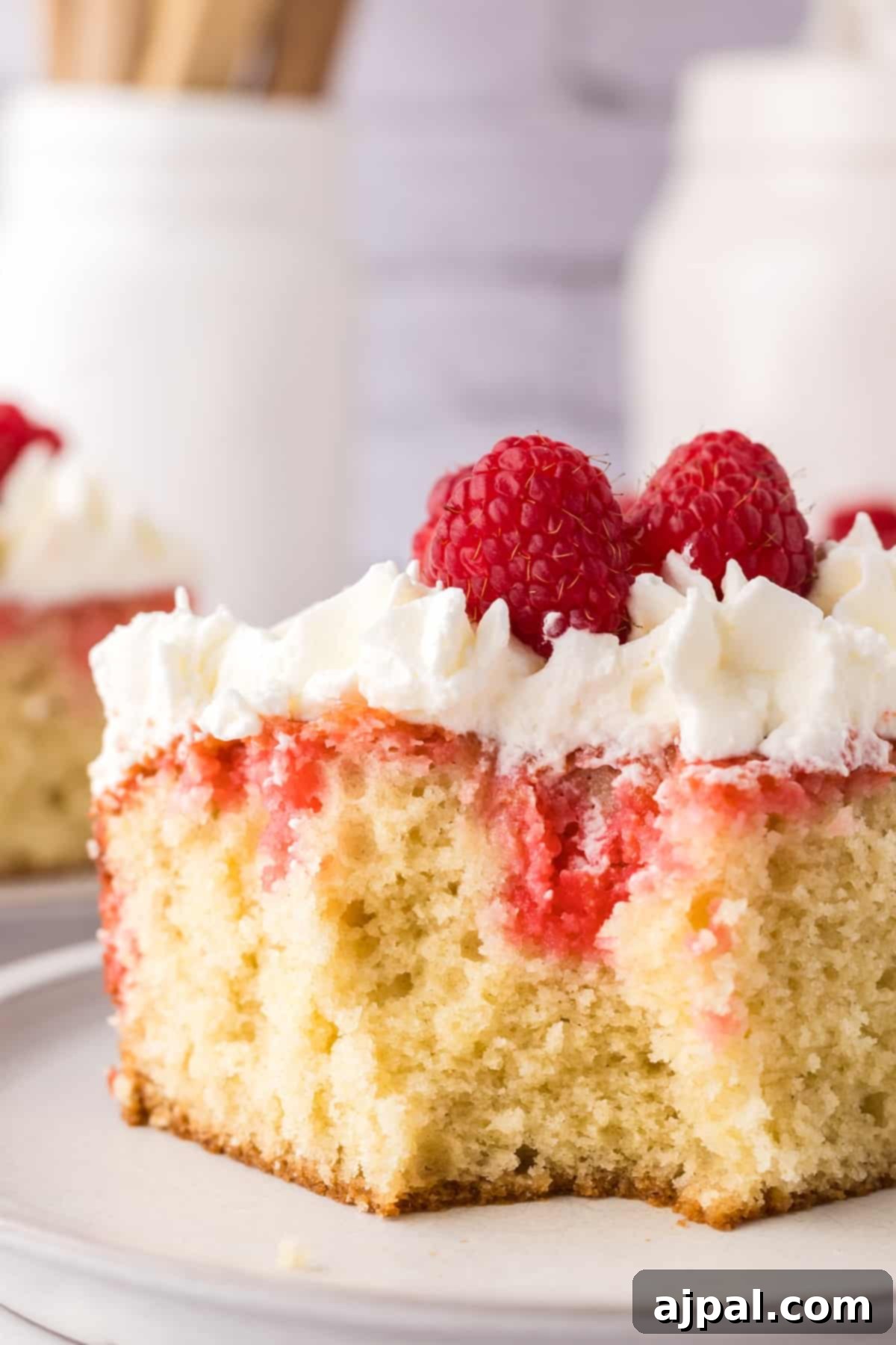 A delightful slice of raspberry sheet cake, beautifully topped with a swirl of whipped cream and garnished with fresh, bright red raspberries, resting on a pristine white plate.
