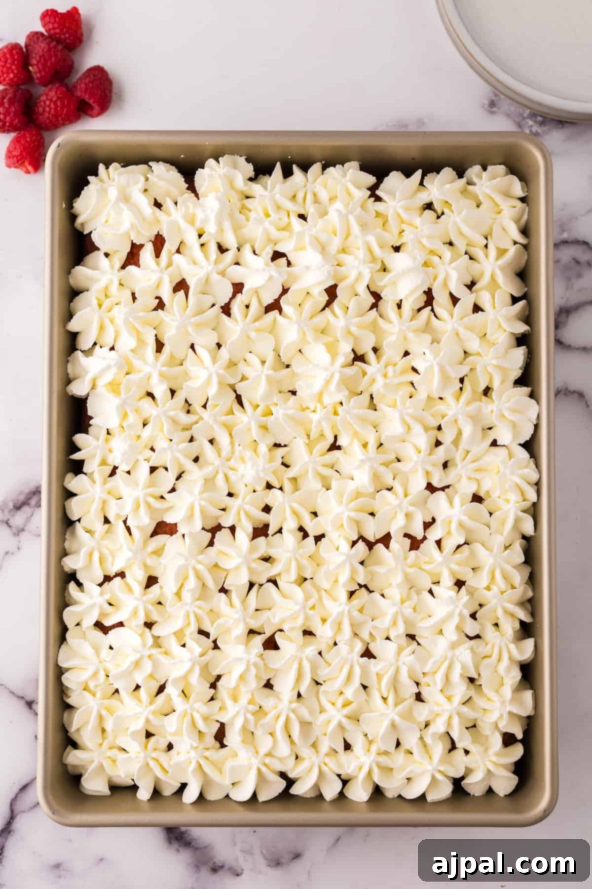 A beautifully frosted sheet cake generously topped with whipped cream and vibrant fresh raspberries, showcasing a luscious texture and inviting appearance, ready to be served.