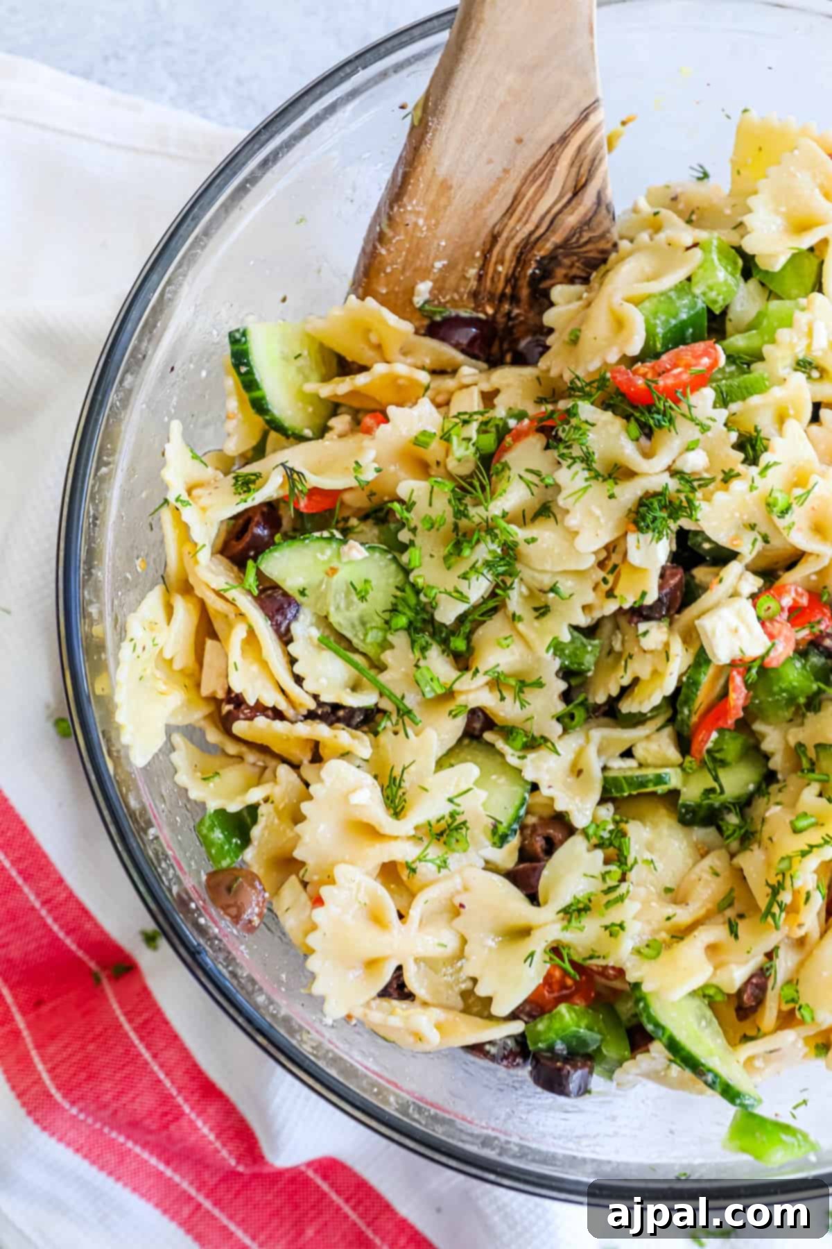 Zesty Greek Pasta Salad 4 A Greek pasta salad in a glass bowl, served with a wooden spoon.
