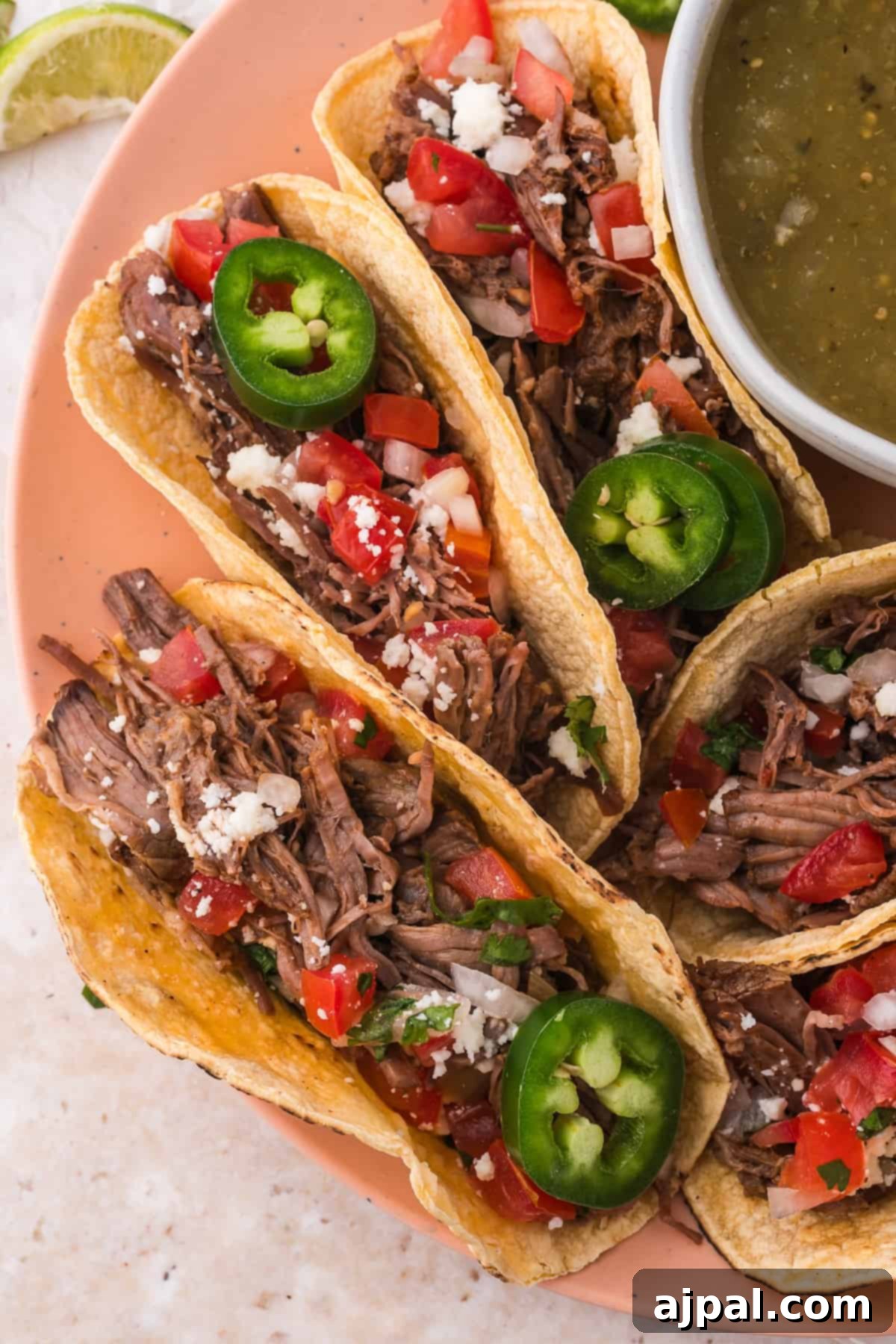 Tender Braised Beef Tacos 2 Plate of shredded beef tacos garnished with jalapeño slices, diced tomatoes, and crumbled cheese with a side of green salsa.