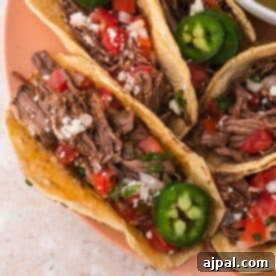 A plate of four shredded beef tacos topped with diced tomatoes, onions, jalapeños, and crumbled cheese.