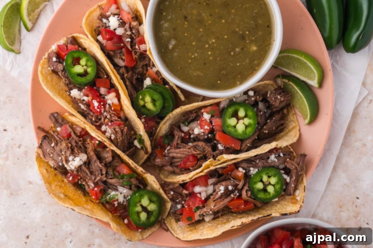 Tender Braised Beef Tacos 3 Three perfectly assembled shredded beef tacos, generously topped with fresh jalapeños, crumbled Cotija cheese, and bright diced tomatoes, artfully arranged with a side of vibrant green salsa and tangy lime wedges.