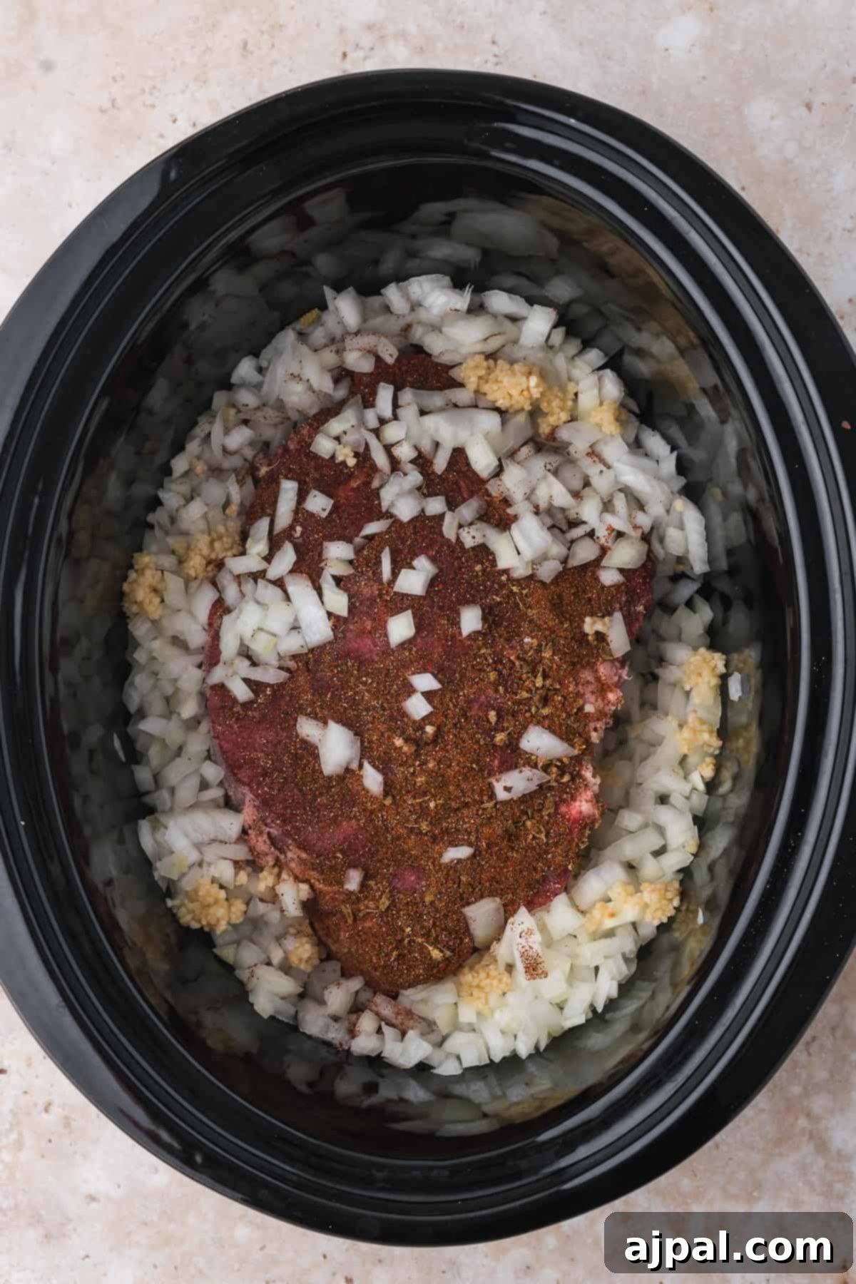 Tender Braised Beef Tacos 7 A raw seasoned beef roast placed on top of chopped onions and garlic in a slow cooker, ready for cooking into shredded beef tacos.