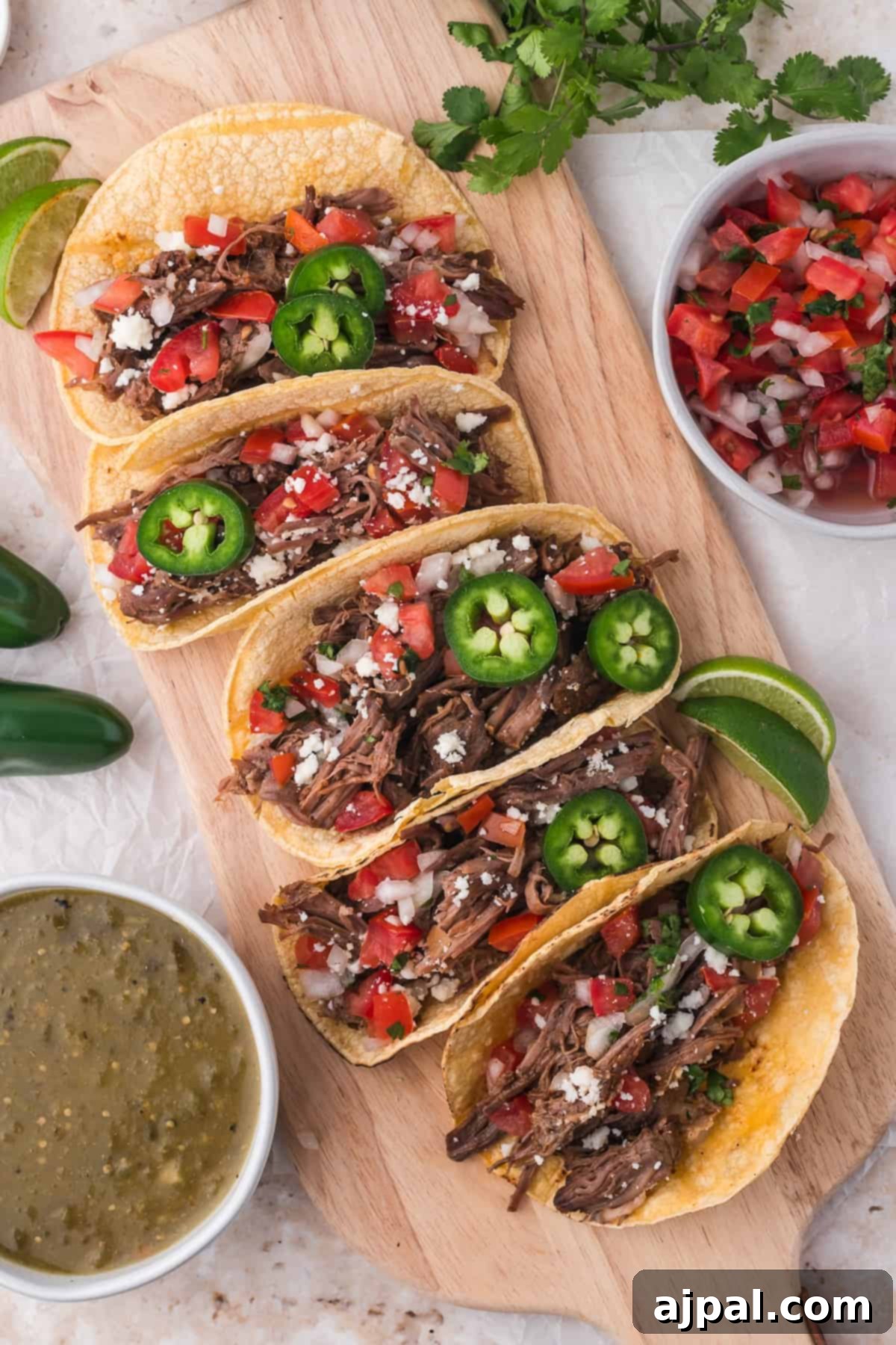 Tender Braised Beef Tacos 10 A wooden board with a selection of beef tacos filled with shredded beef, topped with jalapeños, cheese, and garnished with lime wedges, accompanied by salsa and cilantro.