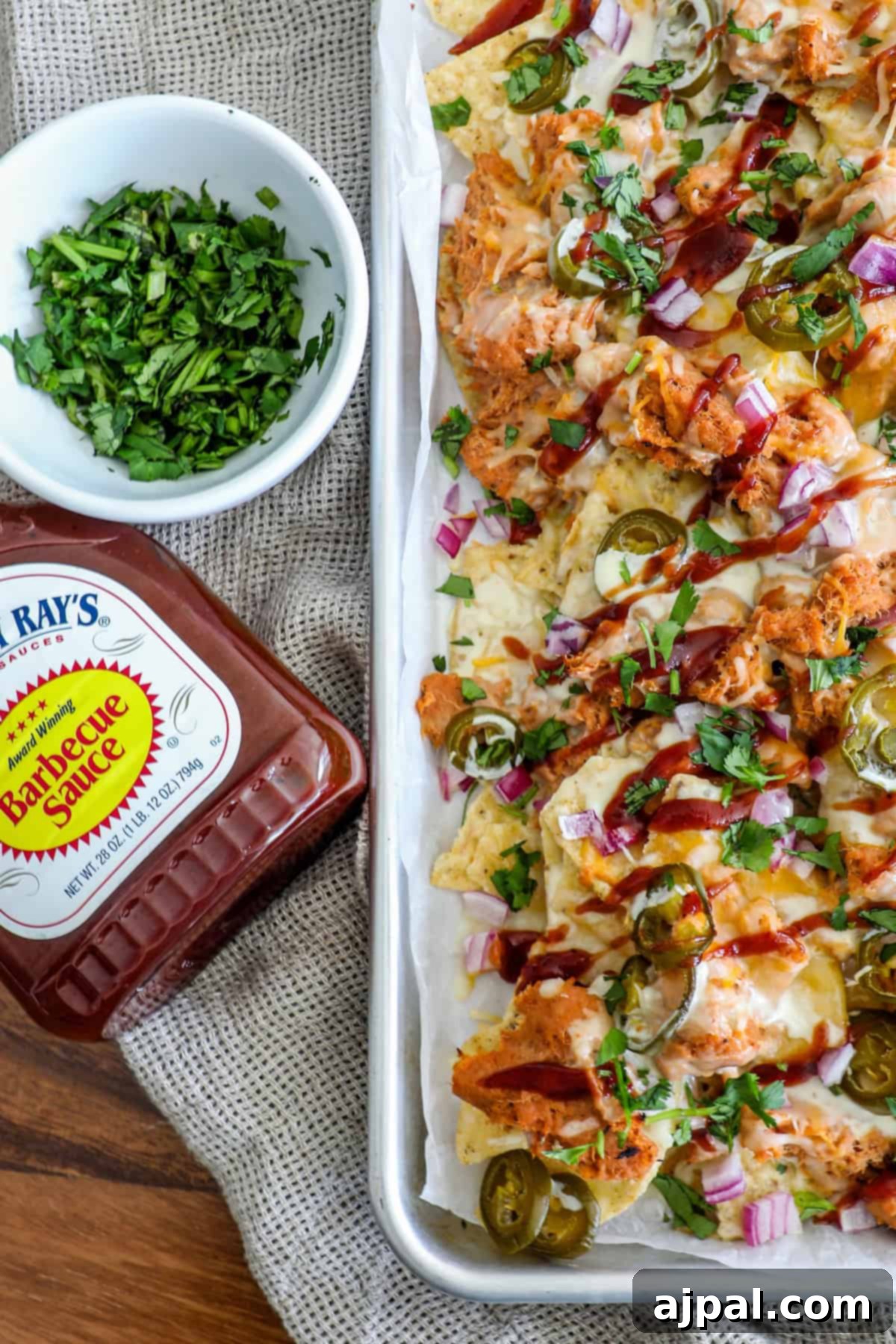 A stunning tray of barbecue chicken nachos, generously topped with vibrant jalapeños, crisp red onions, and fresh cilantro, perfectly complemented by a bottle of rich barbecue sauce.