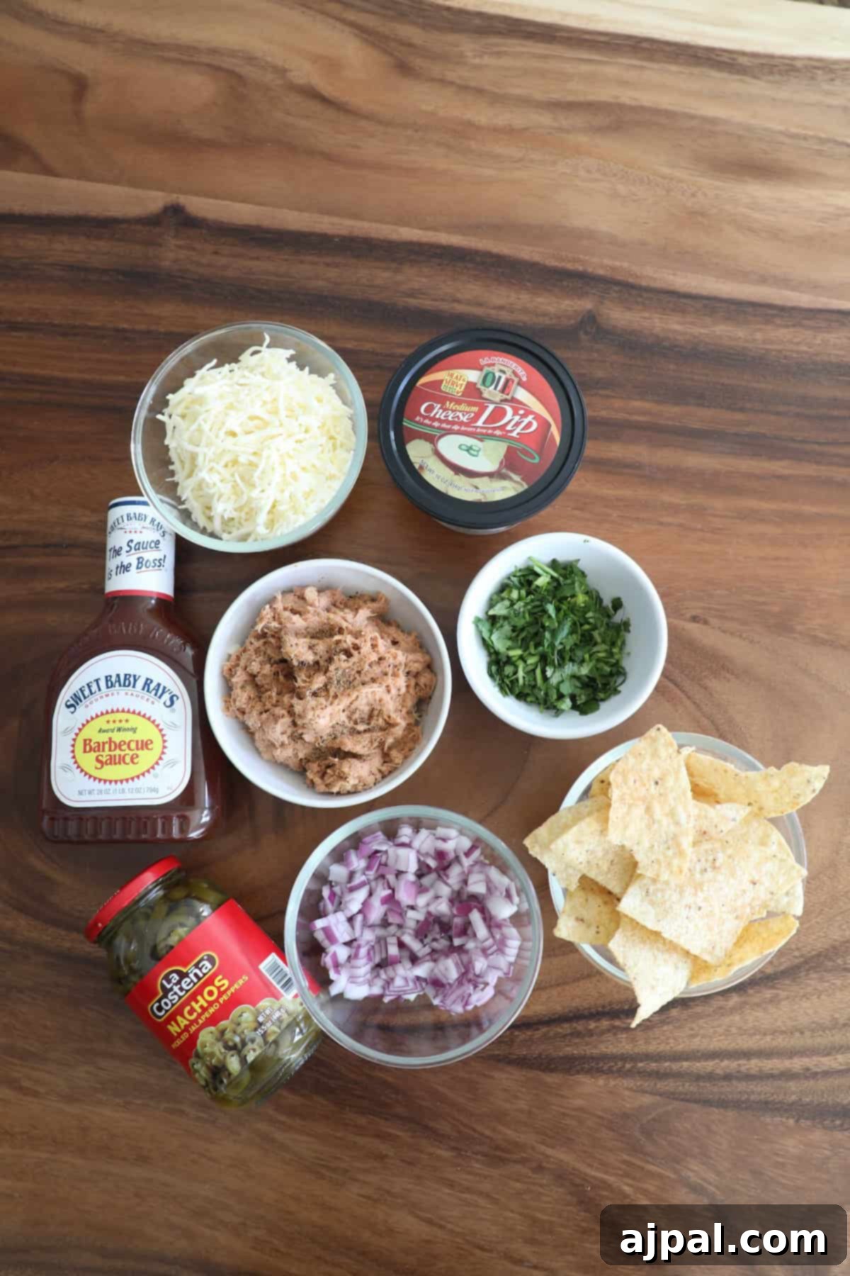Ingredients for a recipe arranged on a wooden table, including shredded cheese, barbecue sauce, canned dip, tuna, chopped onions, jalapeños, cilantro, and tortilla chips.