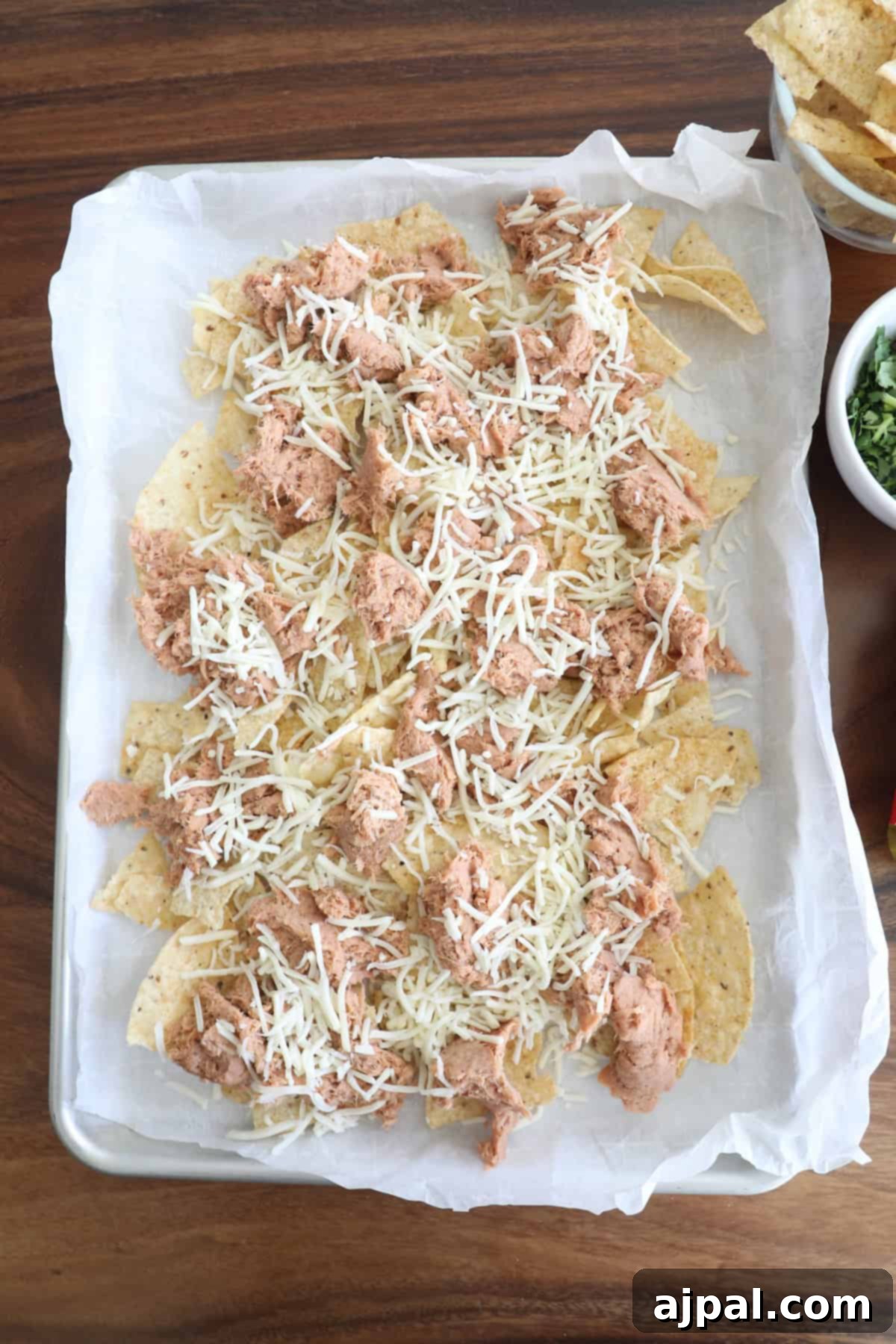 Overhead view of a tray of nachos with shredded cheese and chunks of chicken, placed on a wooden table.