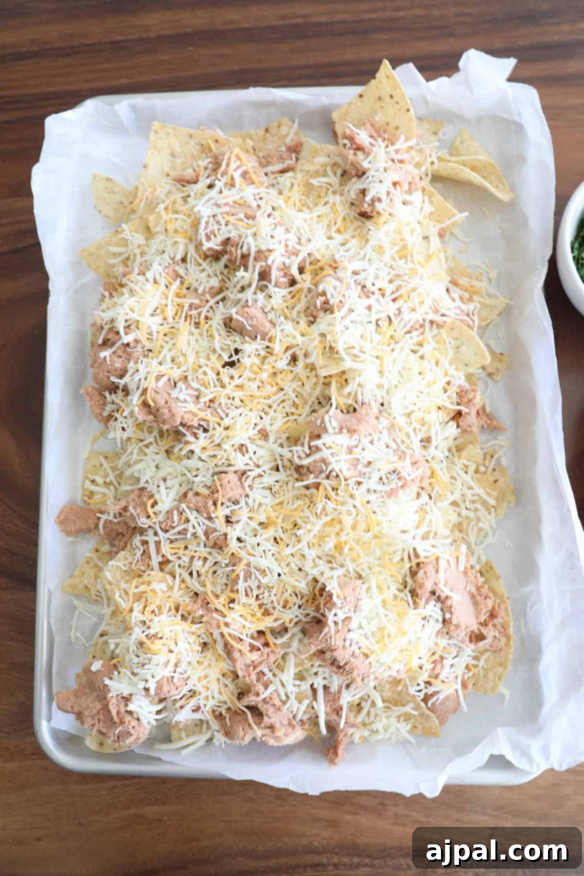 A tray of nachos with shredded cheese and chicken on parchment paper, viewed from above on a wooden table.
