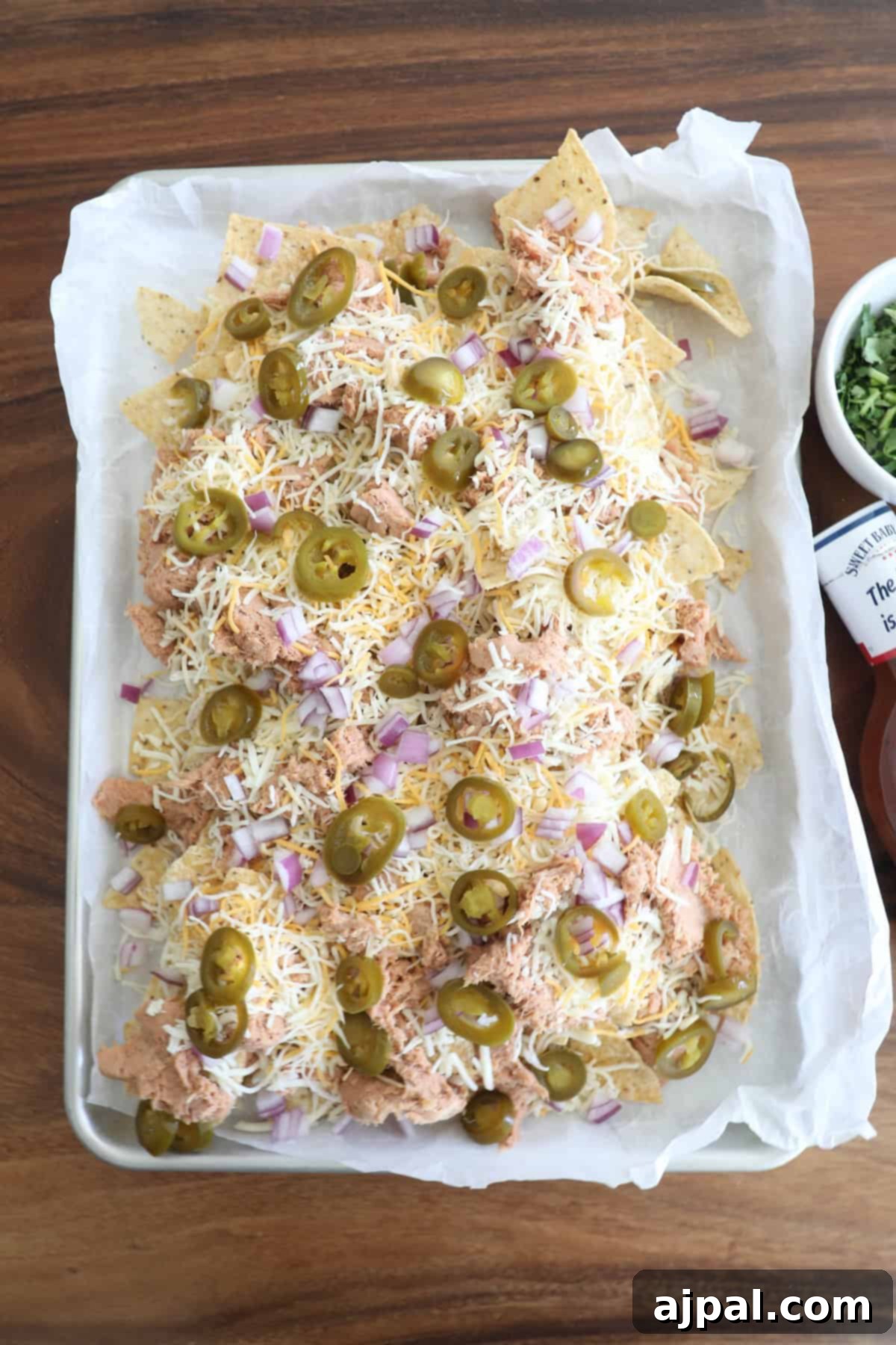 A tray of nachos topped with shredded cheese, jalapeños, red onions, and pink-hued shredded meat, served on wax paper with condiments on the side.