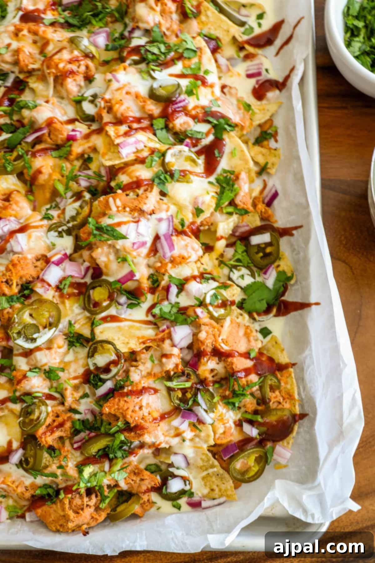 A fully loaded tray of BBQ chicken nachos, adorned with melted cheese, juicy shredded chicken, vibrant jalapeños, crisp red onions, and a delicate drizzle of sauces, artfully presented on a rustic wooden table.
