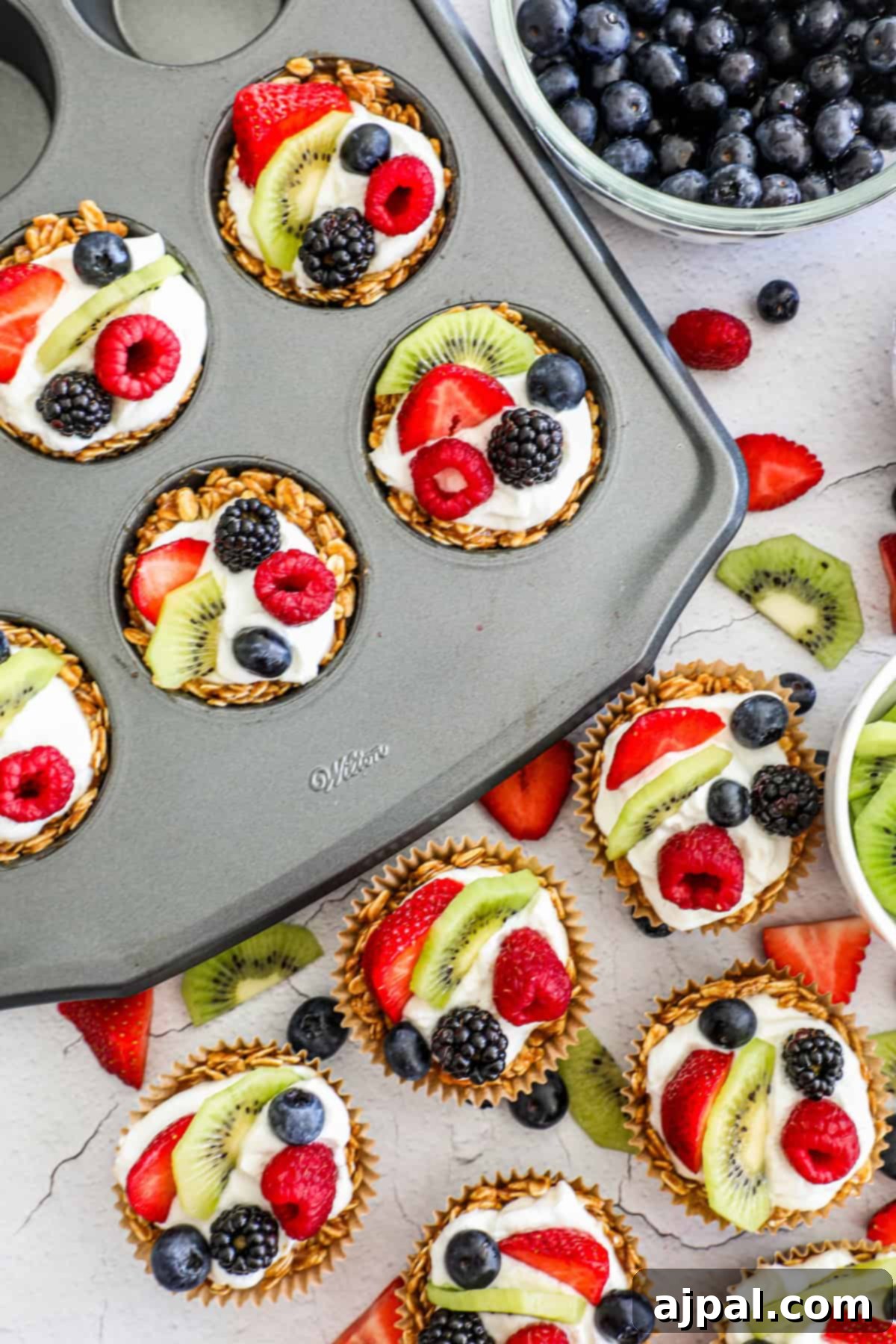 Fruity Granola Bites 3 Two granola fruit cups are presented in a muffin holder, each filled with creamy yogurt and topped with an assortment of fresh berries and kiwi slices.