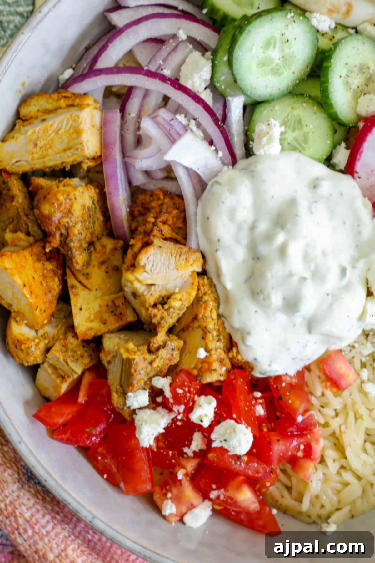 A beautifully arranged plate featuring a Greek chicken gyro bowl with fluffy rice, fresh tomatoes, crisp cucumbers, thinly sliced red onions, a dollop of creamy tzatziki sauce, and crumbled feta cheese.