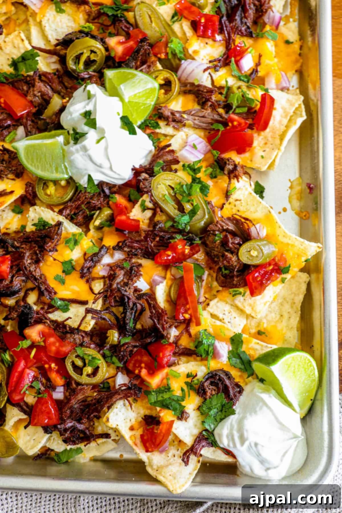 Loaded Carne Asada Nachos 7 A close-up of carne asada nachos on a baking sheet, with various toppings like onions, limes, and sour cream visible.