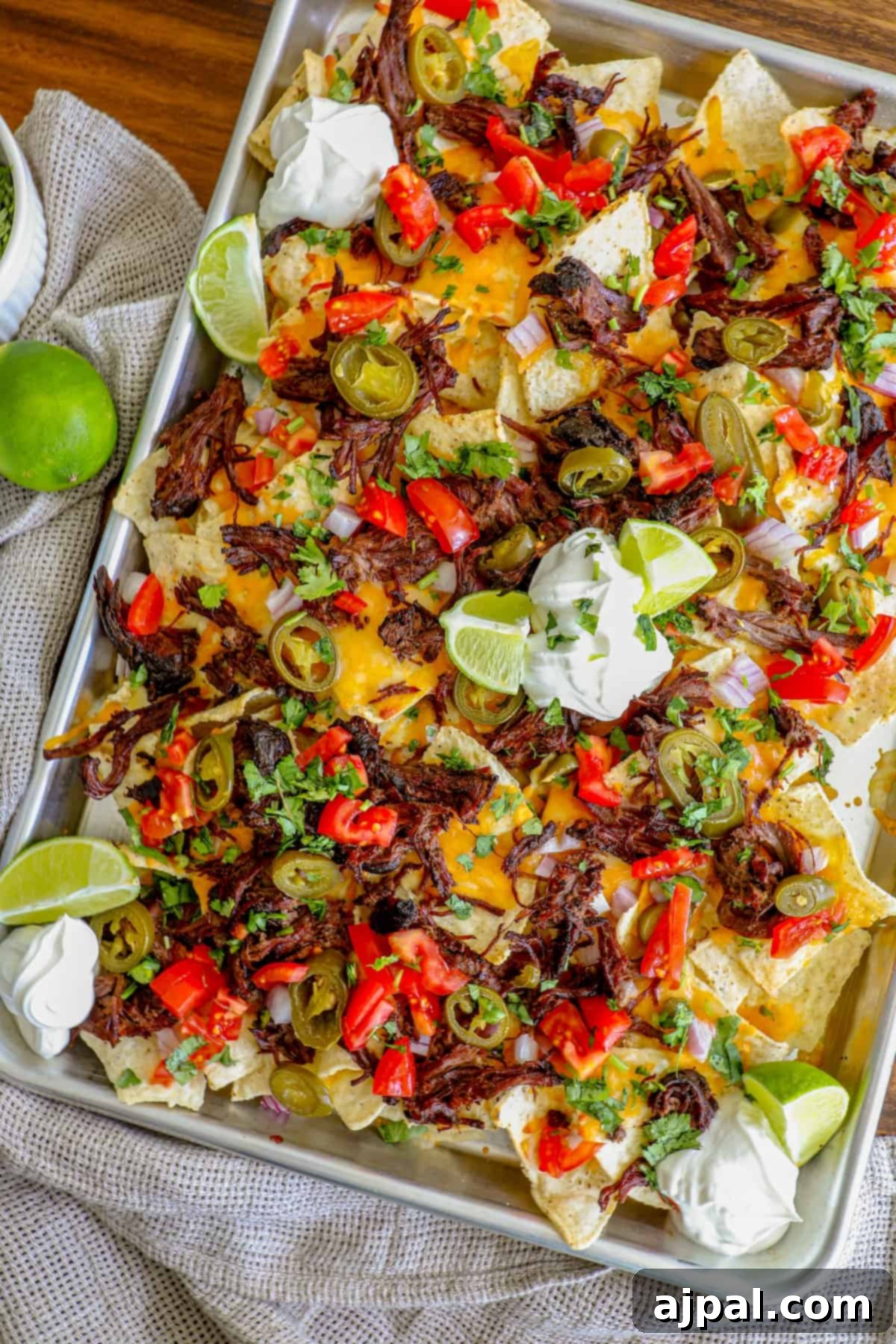 Loaded Carne Asada Nachos 6 A top-down view of a fully loaded tray of carne asada nachos, garnished with chives, jalapenos, onions, lime wedges, and a generous swirl of sour cream.