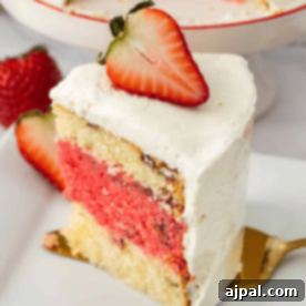 Slice of layered strawberries and cream cake with white frosting, garnished with a halved strawberry on top, positioned on a white plate with a gold fork beside it.