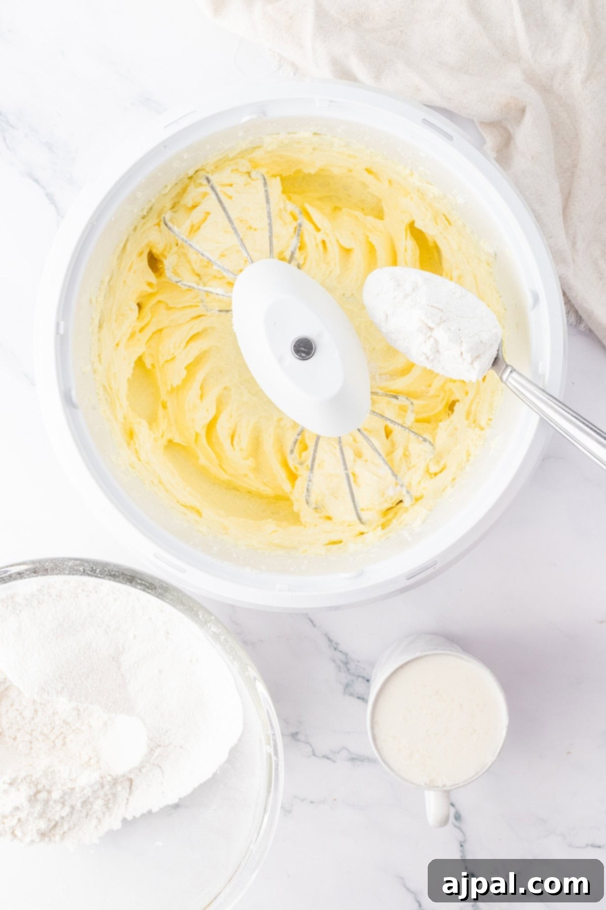 Bowl with whipped butter and flour being added to it, illustrating the cake batter making process.