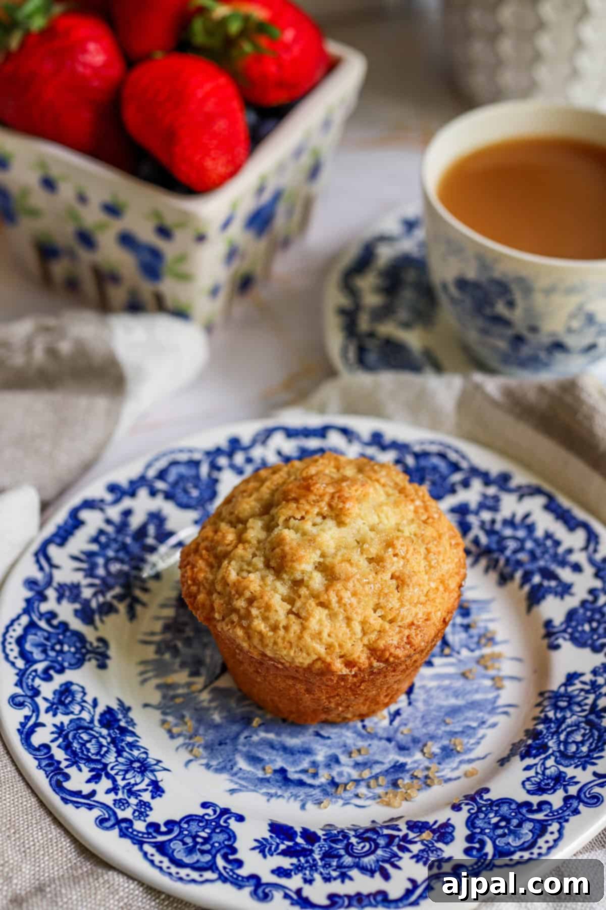 A freshly baked muffin on an ornate blue and white plate, accompanied by a cup of tea and a bowl of strawberries in the background.