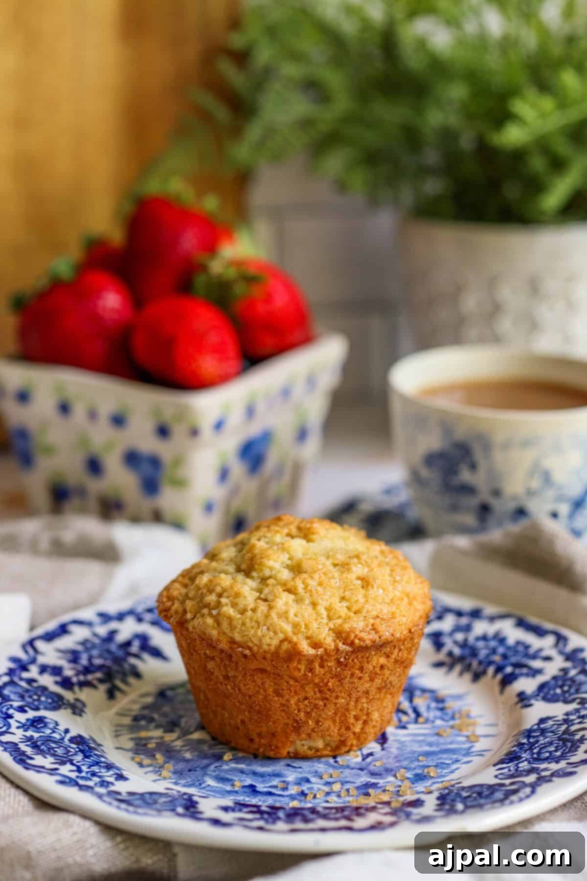 A freshly baked muffin on a blue and white patterned plate, with a bowl of strawberries and a cup of tea in the background.