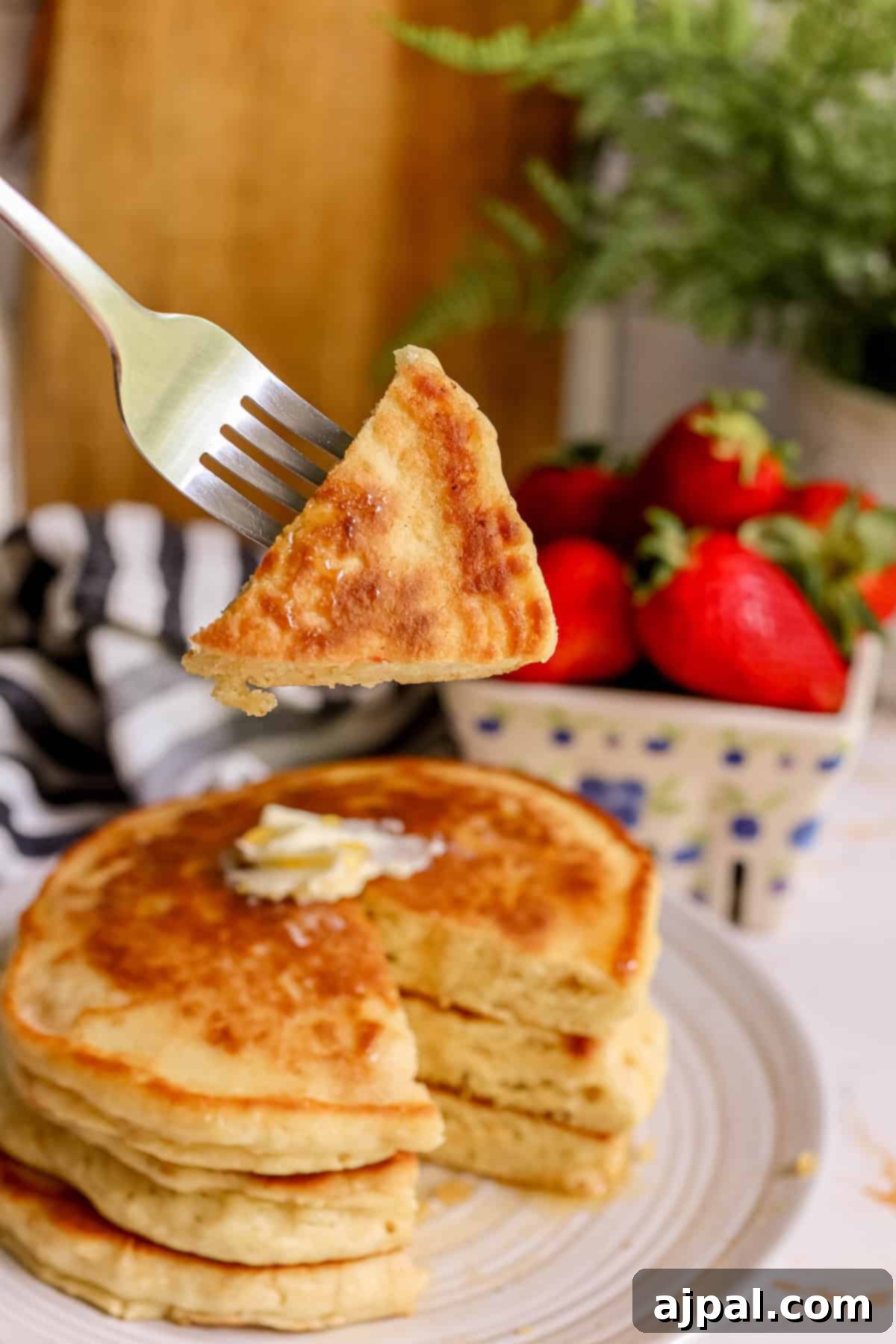 Tangy Sourdough Flapjacks 7 A fork lifts a piece of fluffy sourdough pancake above a stack adorned with butter and syrup, with fresh strawberries invitingly in the soft-focus background.