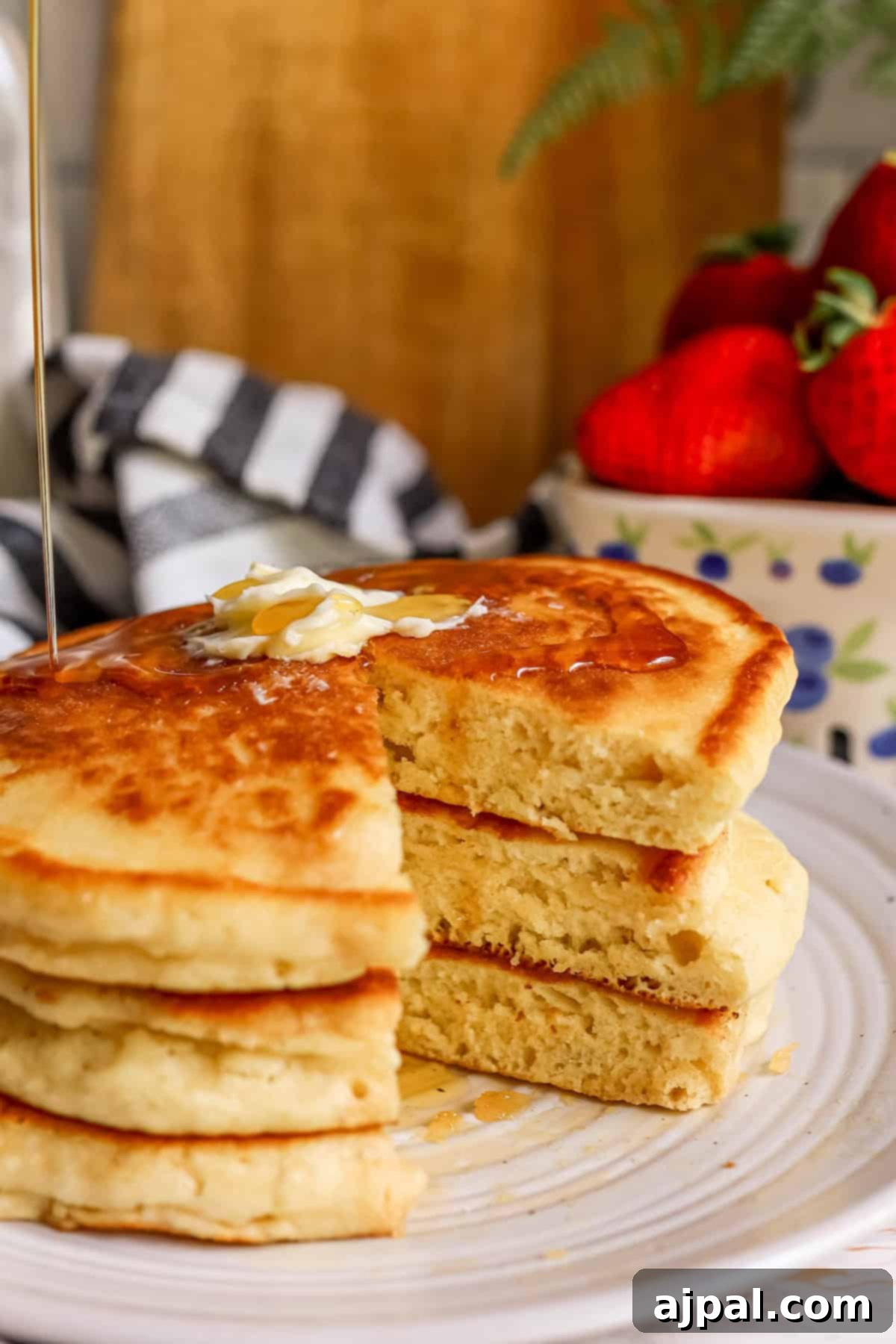 Tangy Sourdough Flapjacks 4 Golden syrup pouring over a stack of fluffy sourdough pancakes, garnished with sliced almonds and fresh strawberries in the background, creating an enticing breakfast scene.