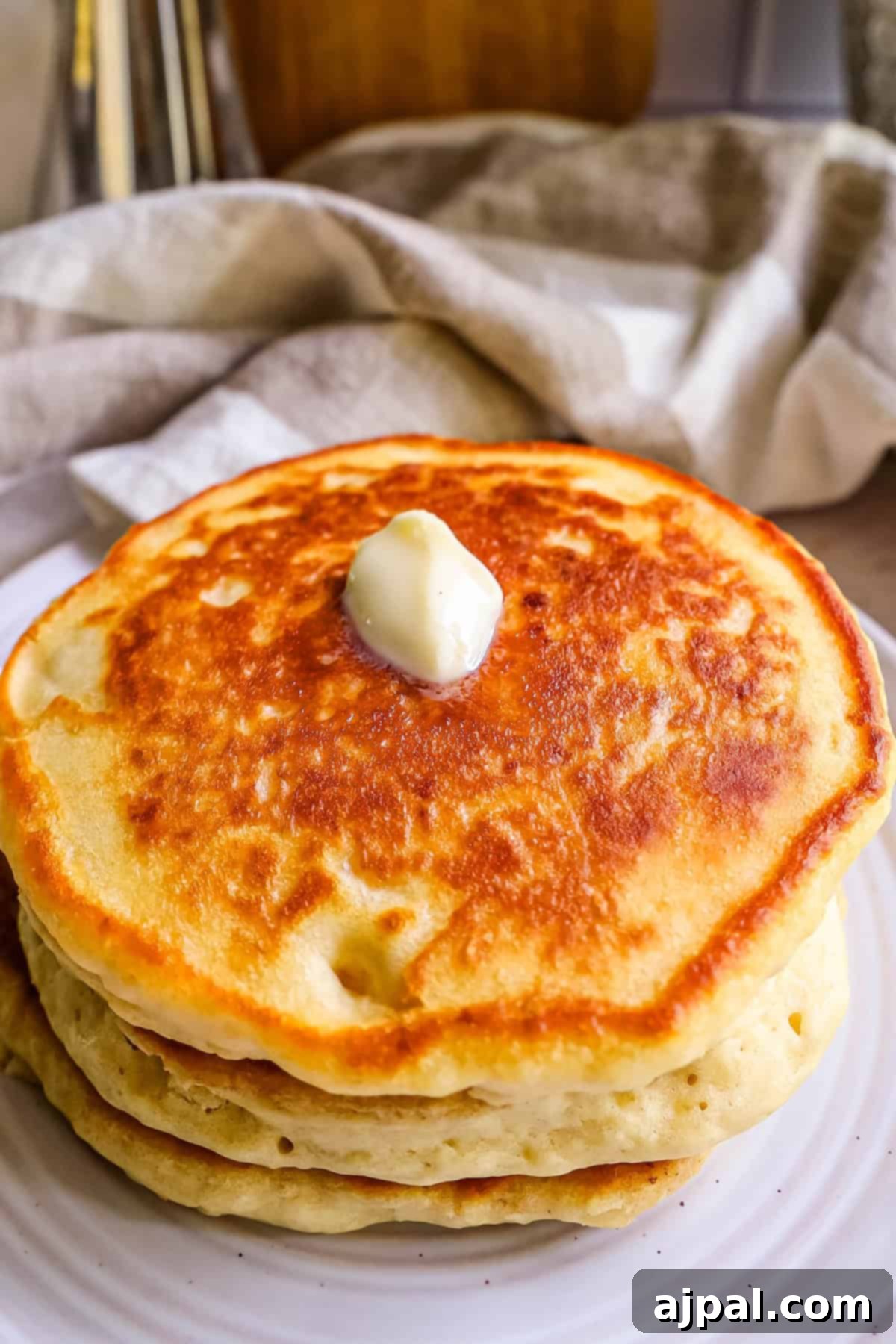 Tangy Sourdough Flapjacks 5 A stack of golden, fluffy pancakes topped with a melting pat of butter, served on a white plate alongside utensils and a napkin, inviting a delicious breakfast.