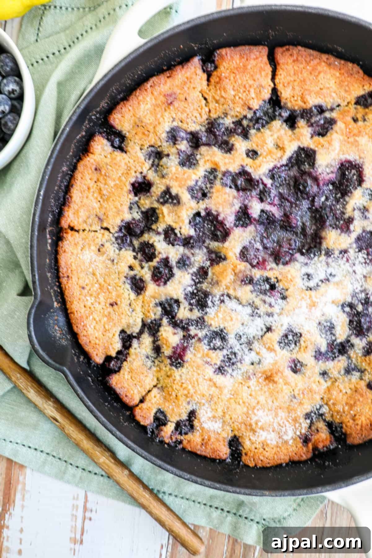 A freshly baked blueberry cobbler in a cast iron skillet, dusted with powdered sugar, on a wooden table with a green cloth.