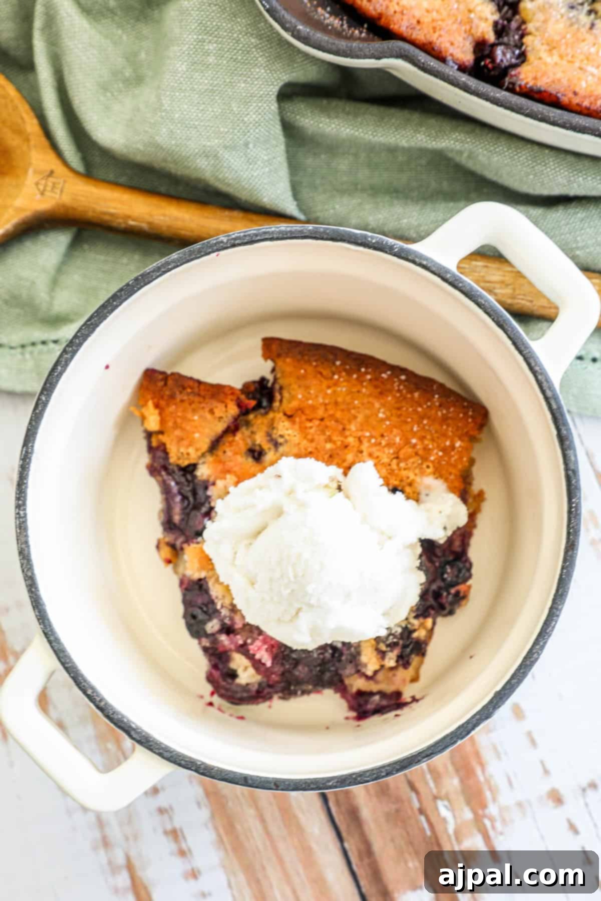 A slice of blueberry cobbler with a scoop of vanilla ice cream in a white ceramic dish, with a rustic wooden background and green cloth.