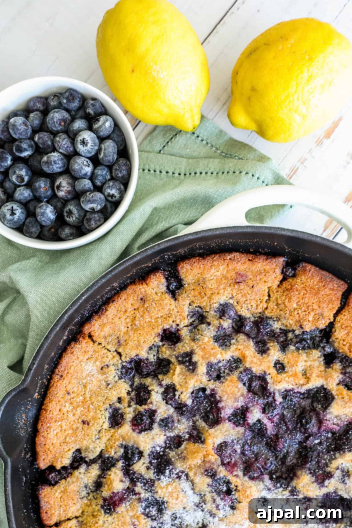 Freshly baked blueberry lemon cake in a skillet beside whole lemons and a bowl of blueberries on a wooden surface with a green napkin.