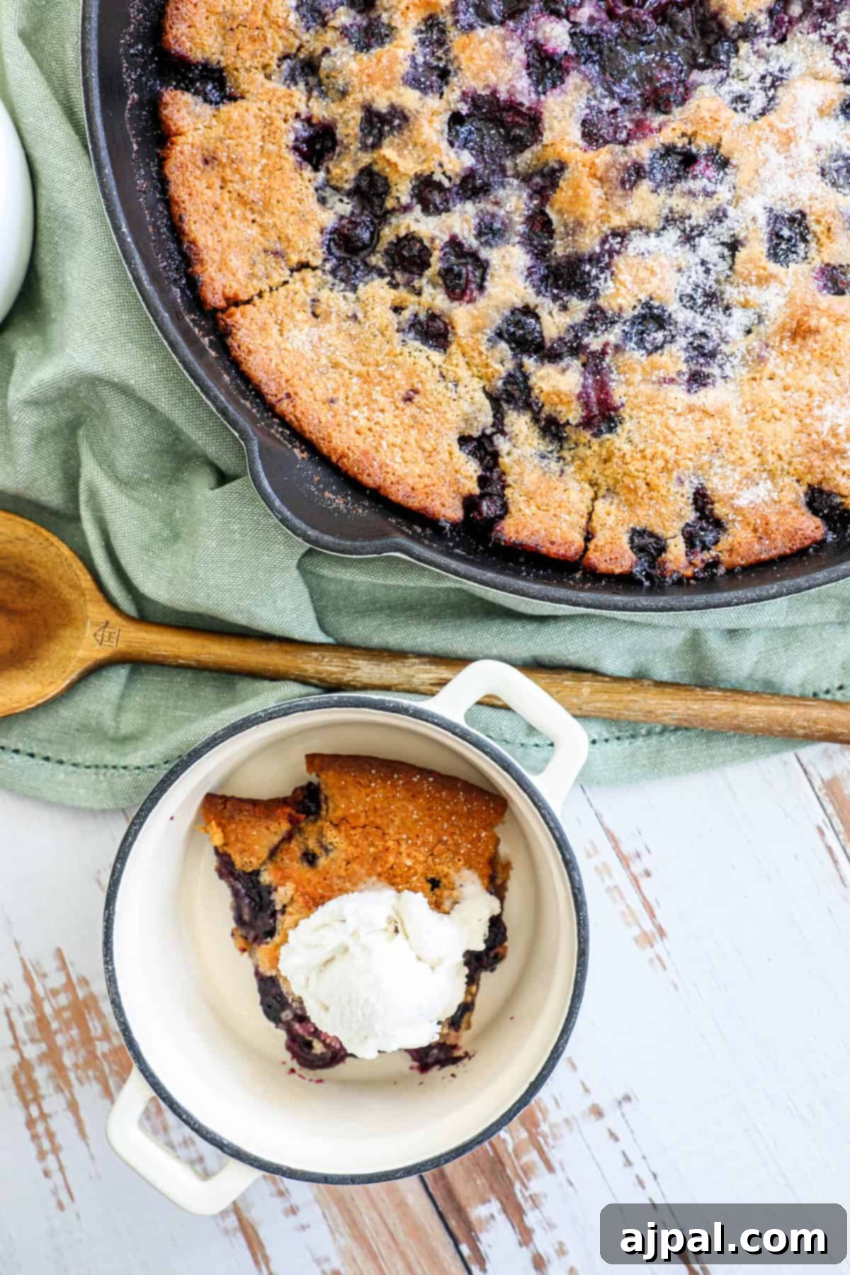 A slice of blueberry skillet cake served with vanilla ice cream in a small bowl, next to the cast iron skillet it was baked in.