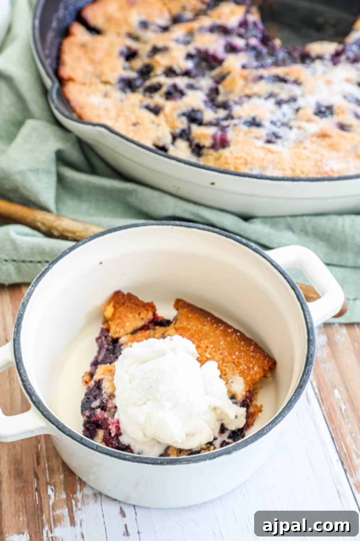 A slice of blueberry cobbler with a scoop of vanilla ice cream in a white enamel dish, with the whole cobbler in the background.