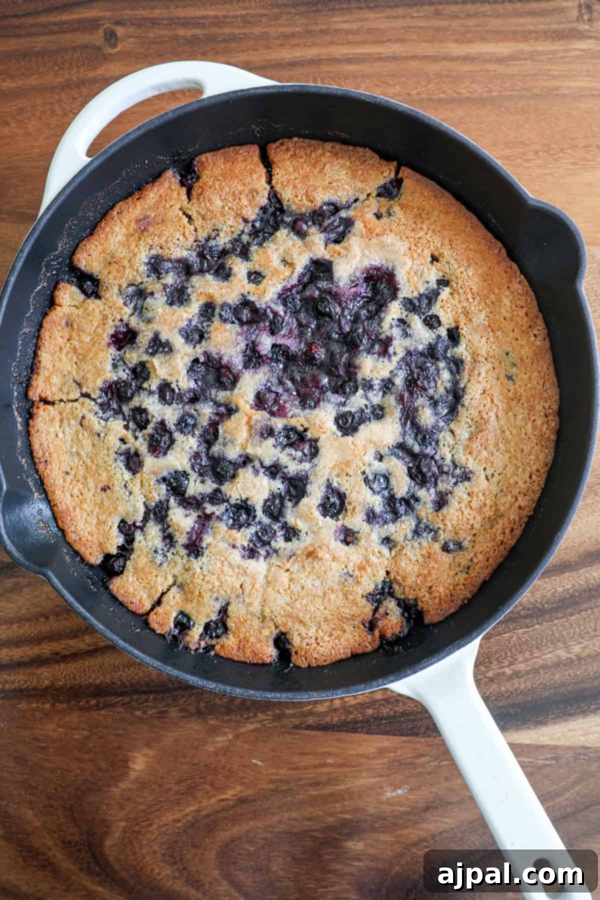 A freshly baked blueberry cake in a white cast iron skillet on a wooden surface.