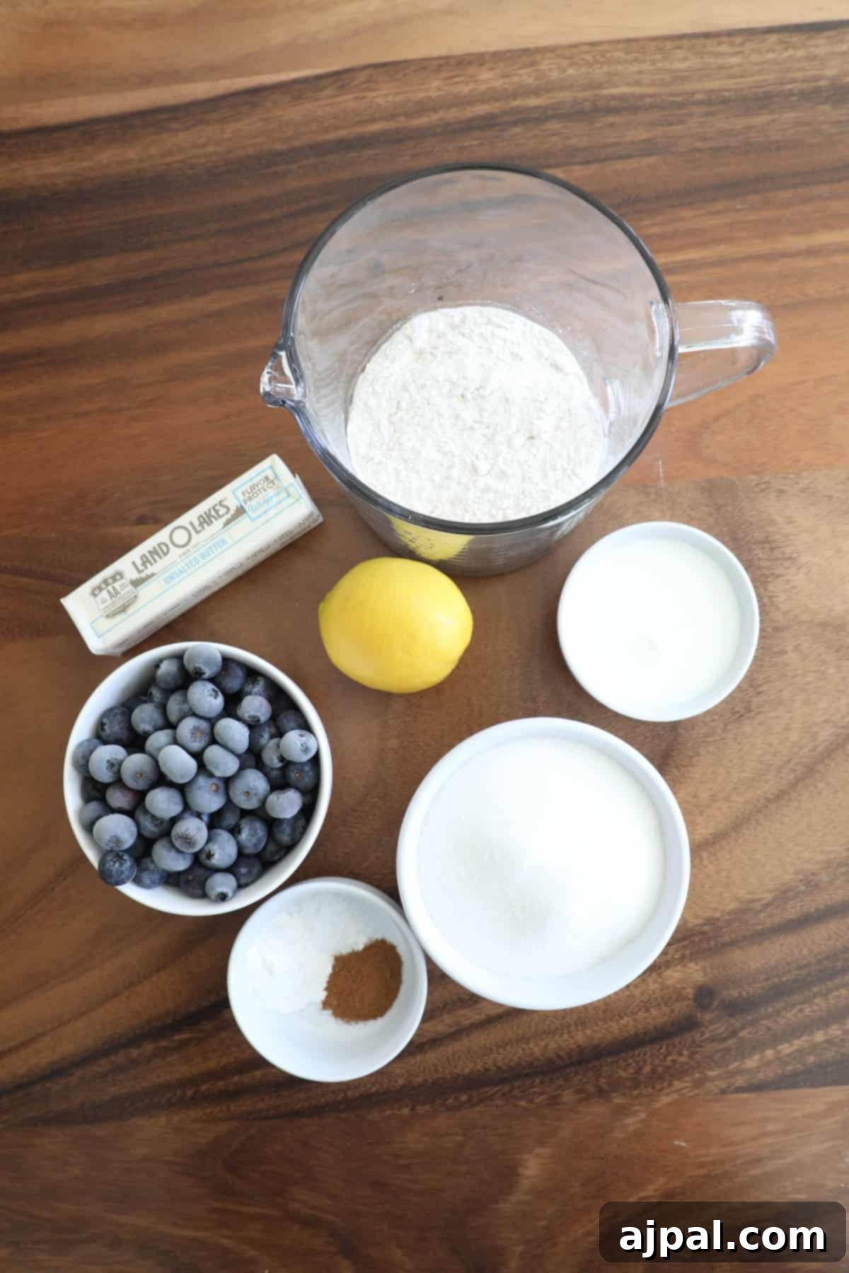 Ingredients for baking arranged on a wooden table, including a measuring cup with flour, blueberries, sugar, milk, butter, a lemon, and a small bowl of baking powder.
