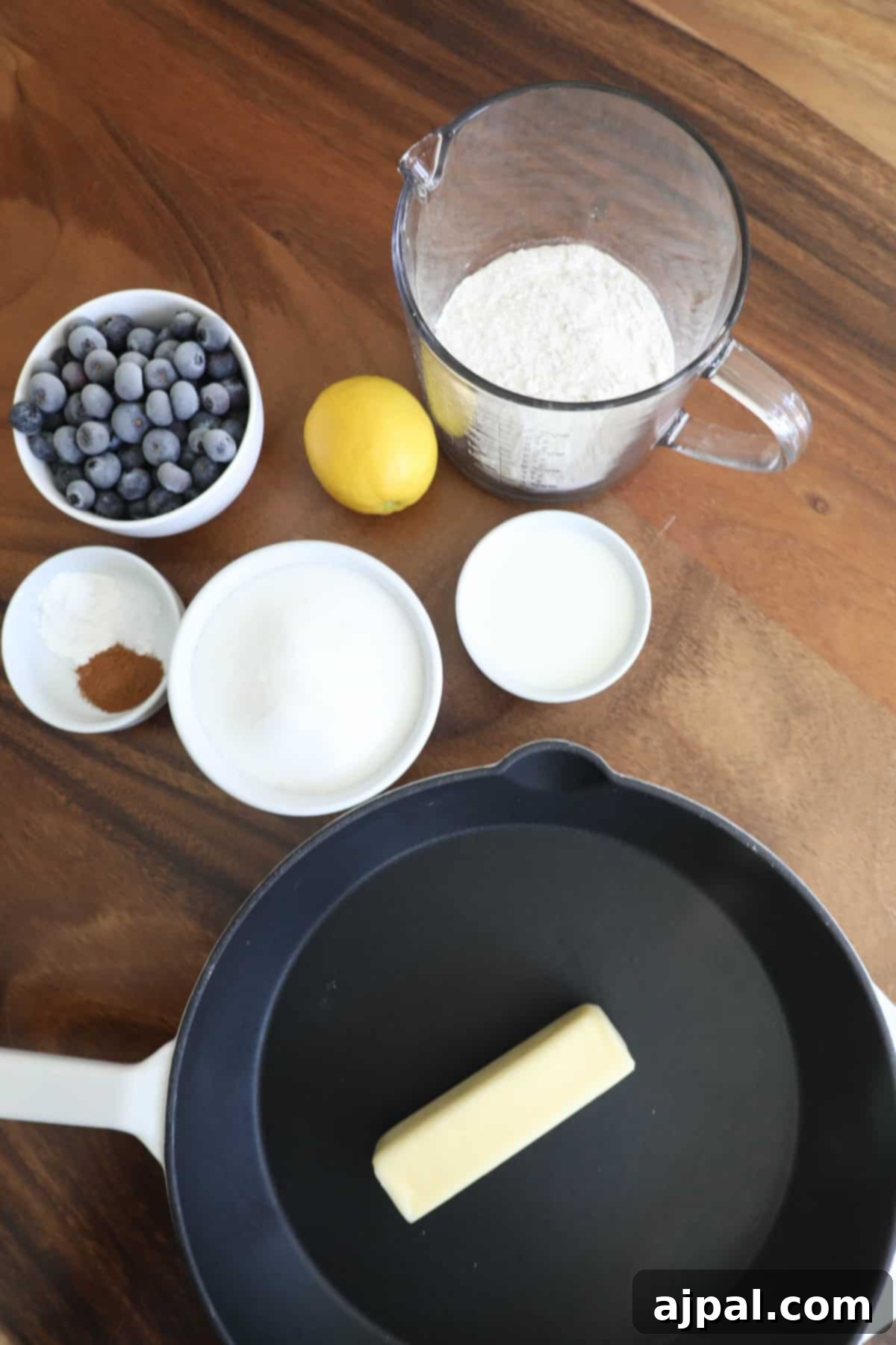Ingredients for making pancakes arranged on a wood table, including flour, blueberries, lemon, sugar, milk, and a skillet with butter.