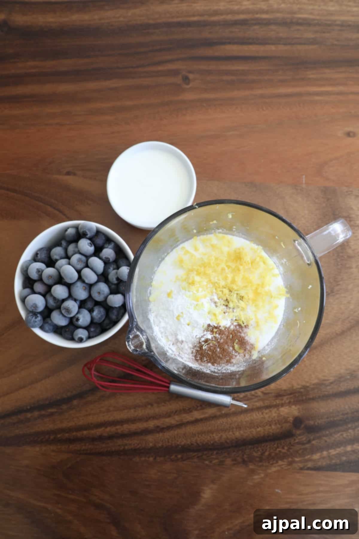 Top-down view of a kitchen counter with ingredients for baking: a bowl of blueberries, a glass of milk, and a mixing bowl with flour, sugar, and grated lemon zest.