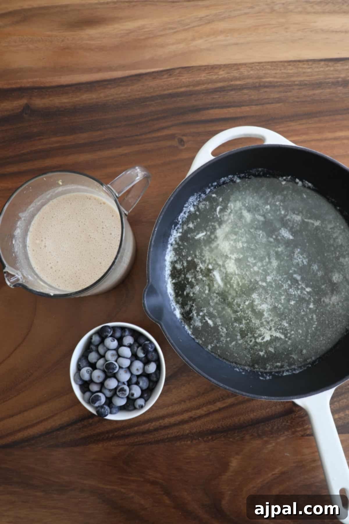 A top view of a kitchen scene with a jug of batter, a skillet with melted butter, and a bowl of blueberries on a wooden countertop.