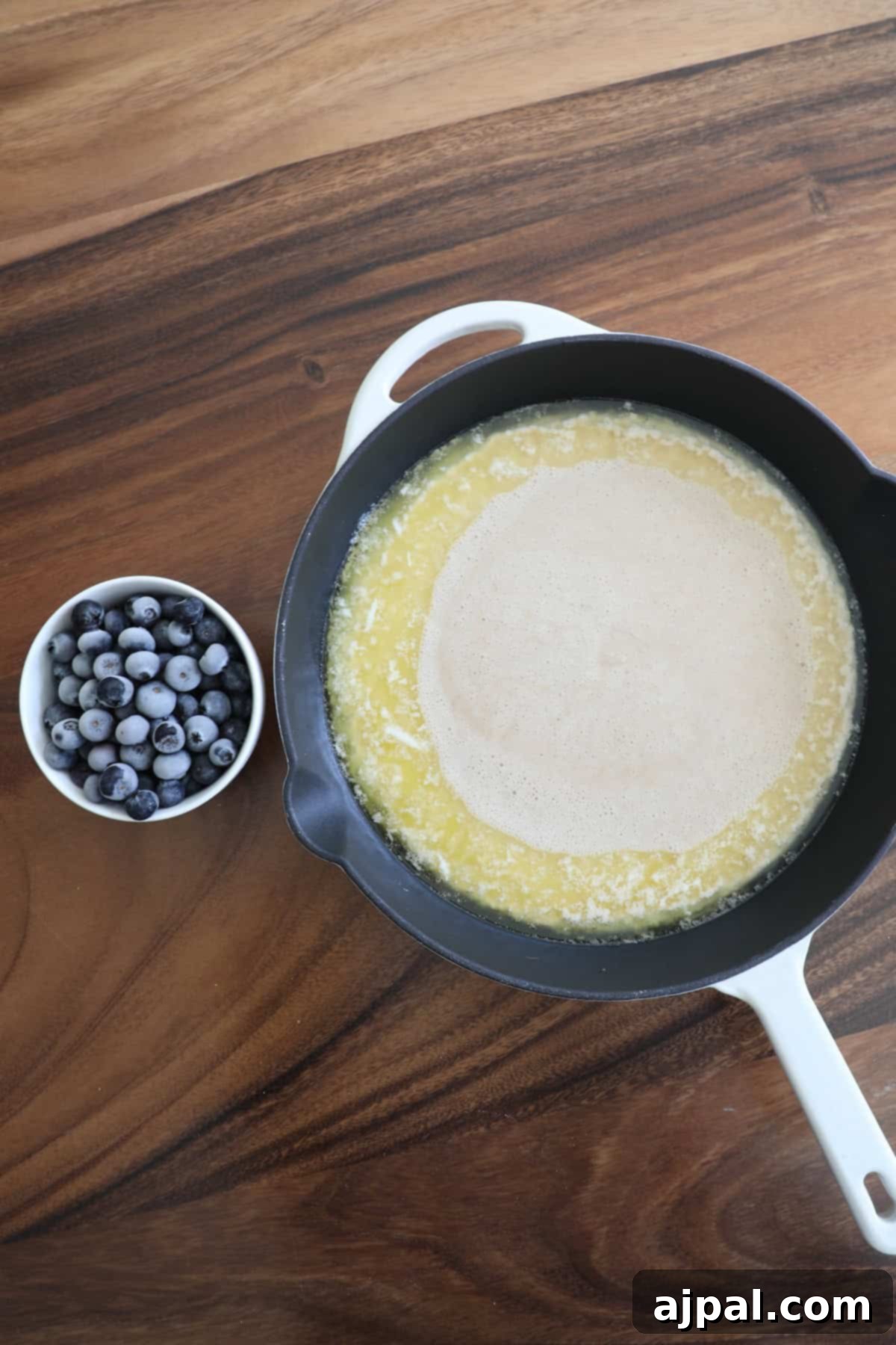 A pancake cooking in a cast iron skillet next to a small bowl of blueberries on a wooden table.