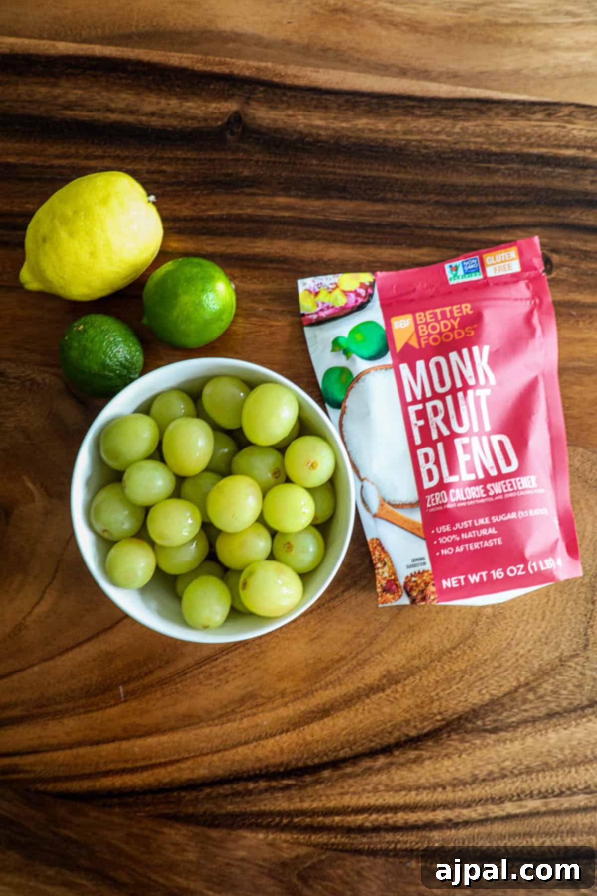 Zingy Sweet Grapes 5 A bowl of green grapes with a lemon, lime, and a bag of monk fruit blend sweetener on a wooden surface.
