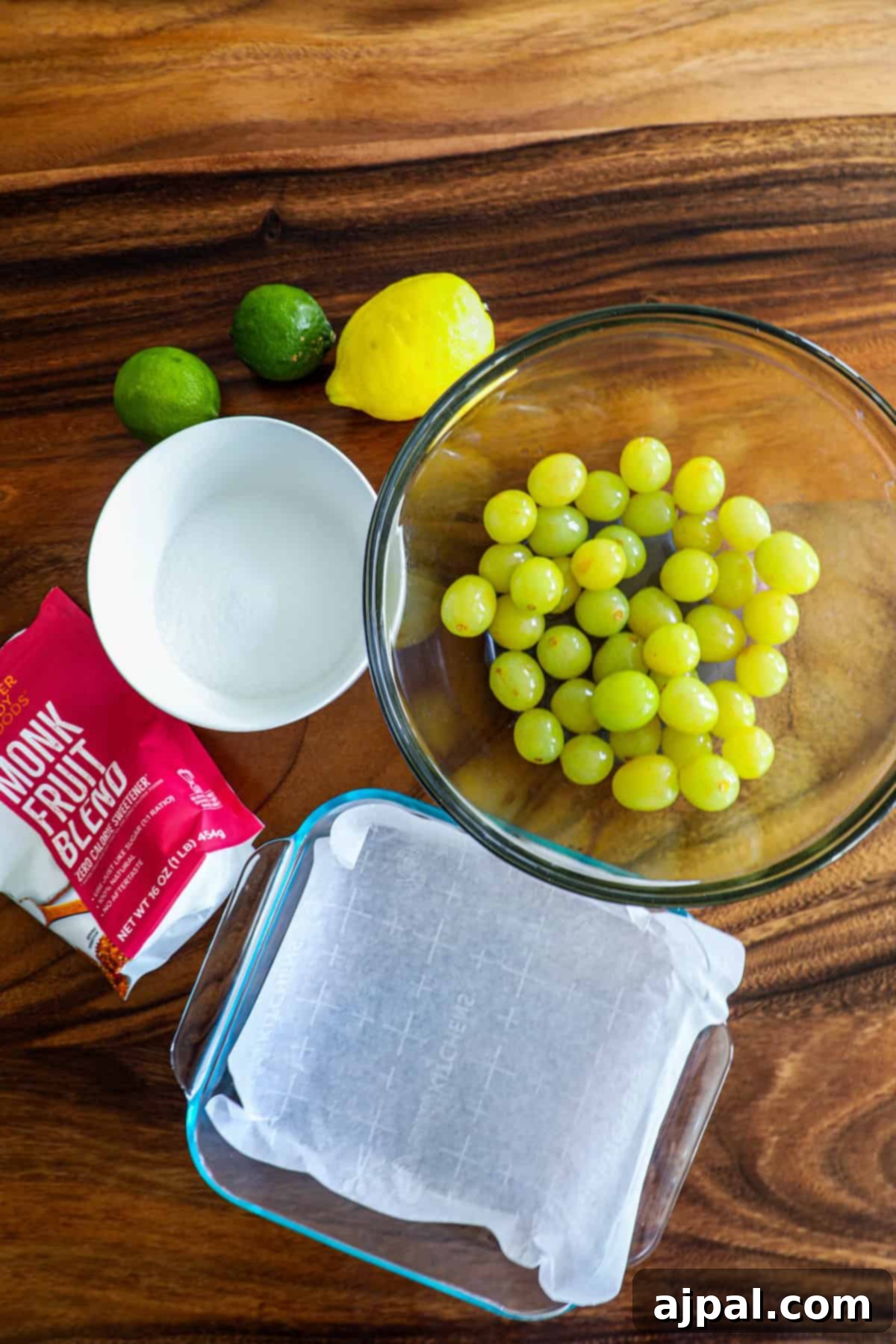 Zingy Sweet Grapes 6 Glass bowl of grapes on a wooden table with lemons, limes, sugar, and a baking dish lined with parchment paper.