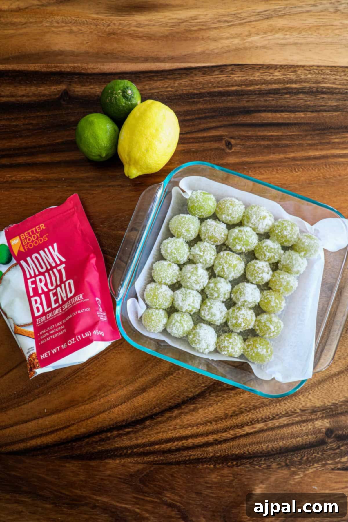 Zingy Sweet Grapes 7 Glass dish of homemade matcha energy balls coated in monk fruit sweetener, alongside a bag of the sweetener and fresh lemons and limes on a wooden surface.