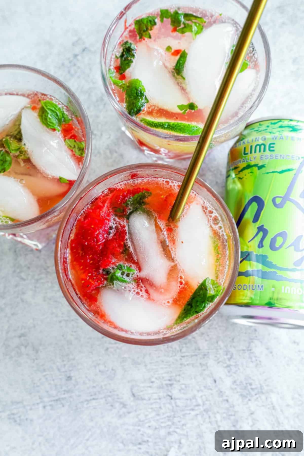 Three glasses of strawberry and lime mocktails with fresh mint leaves and ice, accompanied by a can of lime-flavored sparkling water, on a light textured surface, highlighting the ingredients.