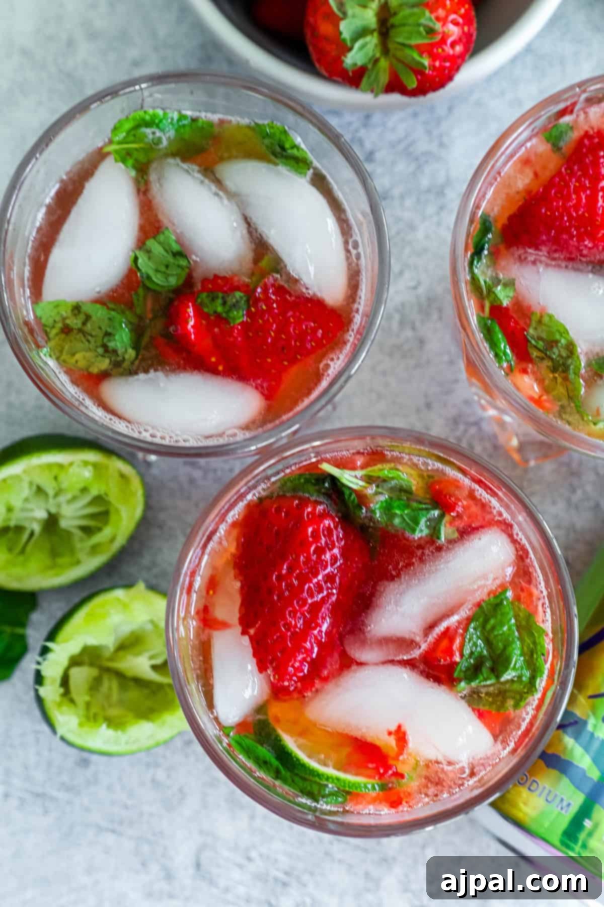 Three glasses of strawberry mojito mocktails with mint leaves and ice cubes on a textured gray surface, viewed from above, showcasing their vibrant color.