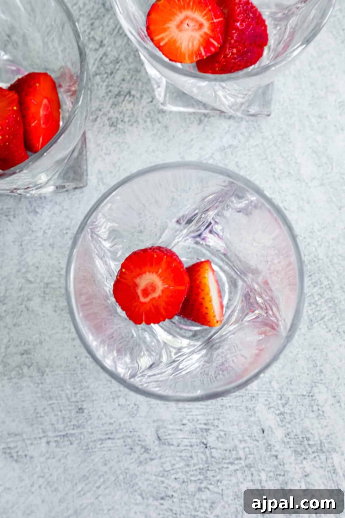 Top view of a clear glass containing water and sliced strawberries, with another similar glass visible in the background on a textured surface, illustrating the preparation process.